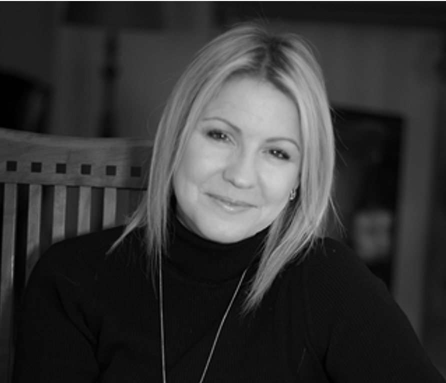 A woman with shoulder-length blonde hair, wearing a black turtleneck, smiling while sitting on a wooden chair indoors.