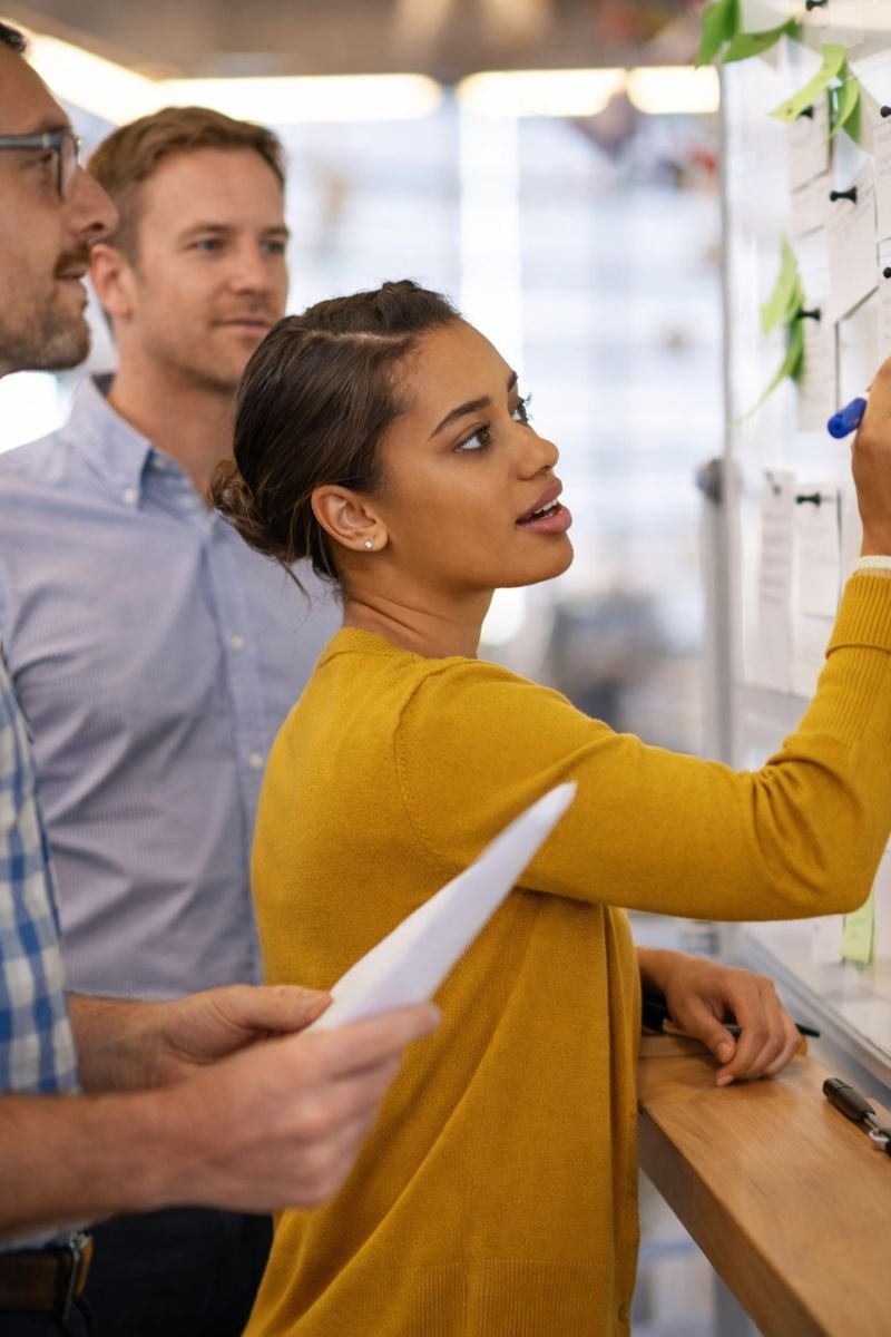 A diverse group of three colleagues working together at a whiteboard, with one woman in a yellow top writing notes and two men observing.