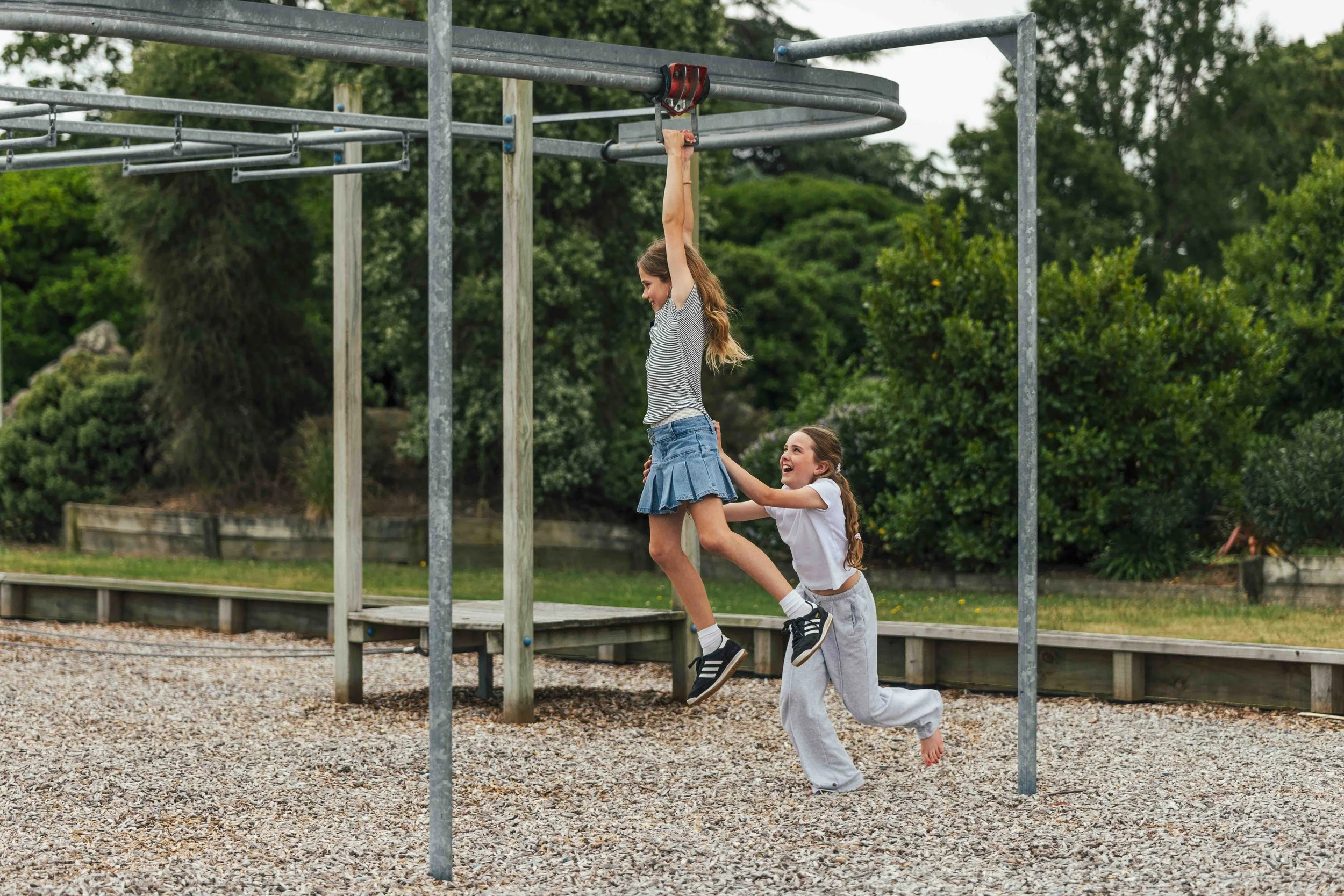 Two girls playing on a playground, one hanging from a metal bar while the other pushes her from below, surrounded by green trees and gravel ground.