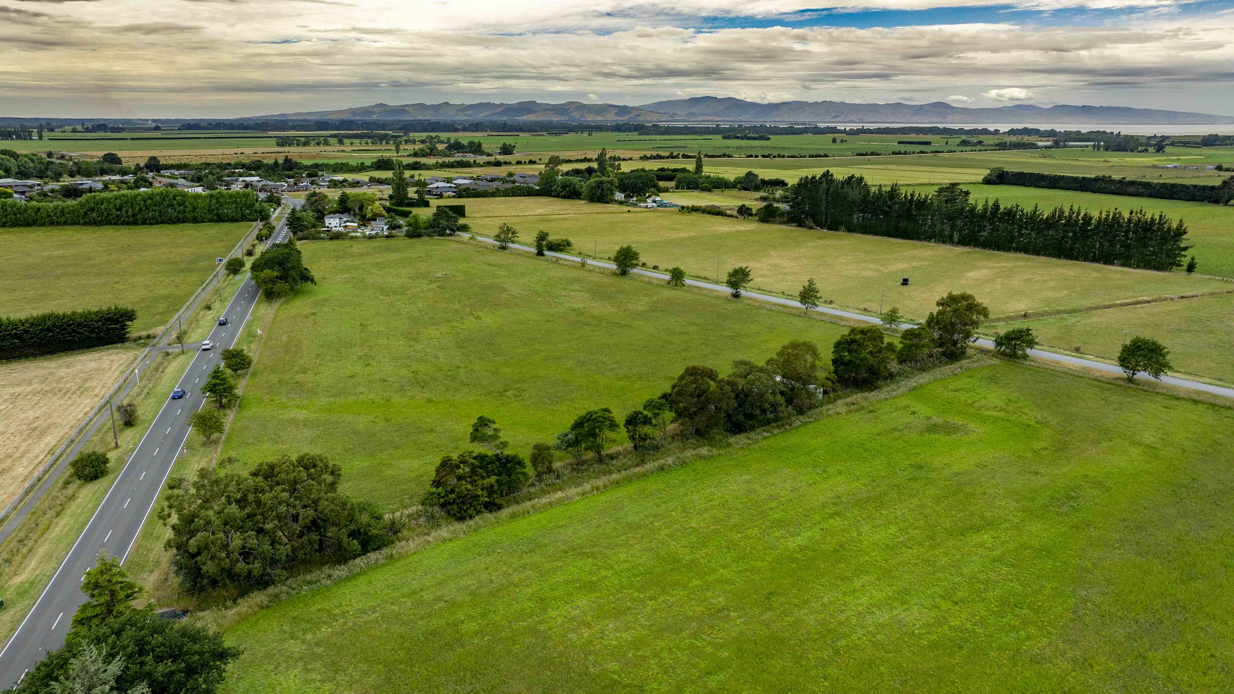 Aerial view of green farmland with a two-lane road running through it, houses in the background, and distant mountains under cloudy skies.