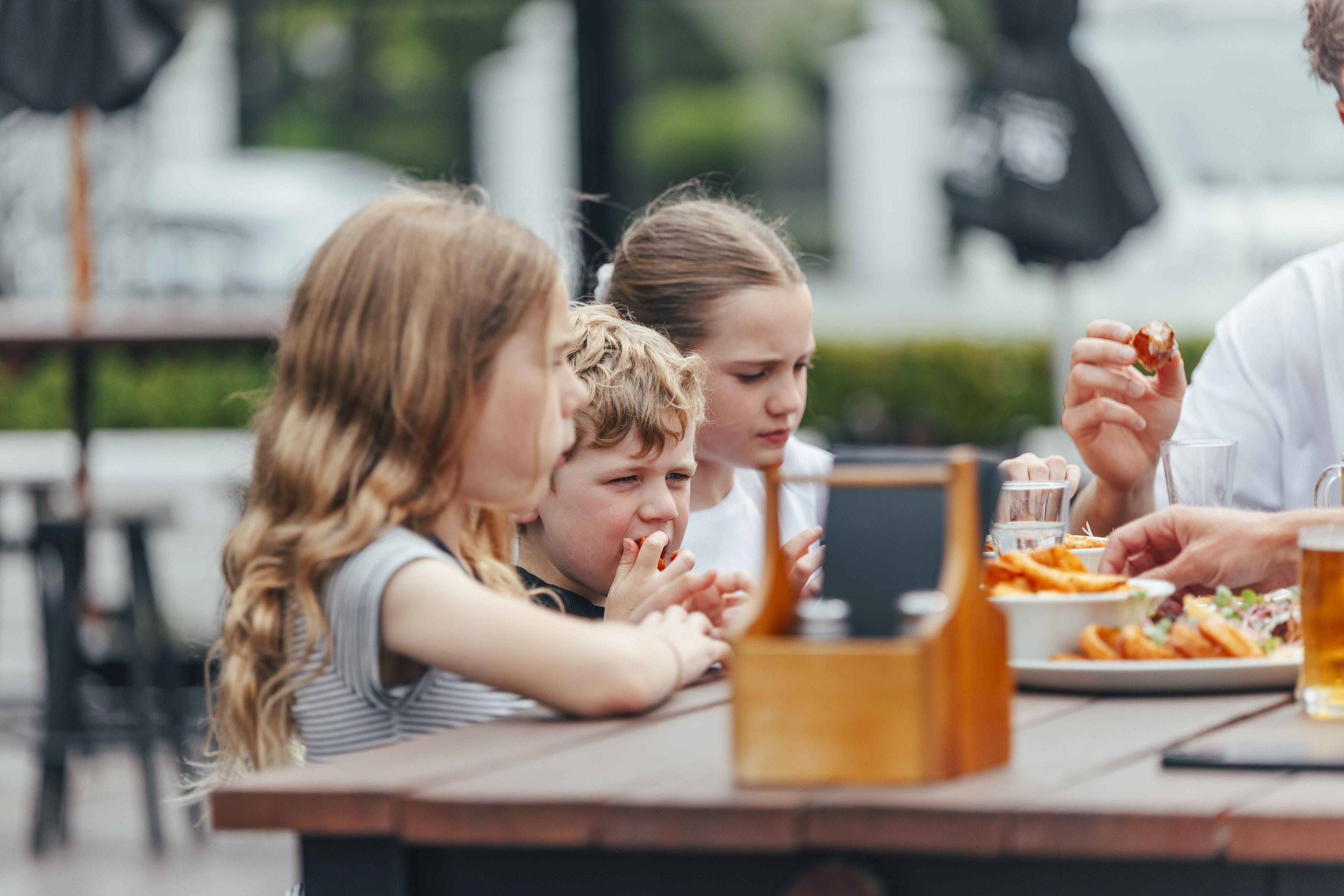 Children sitting at an outdoor table eating food, with an adult handing out a dish.
