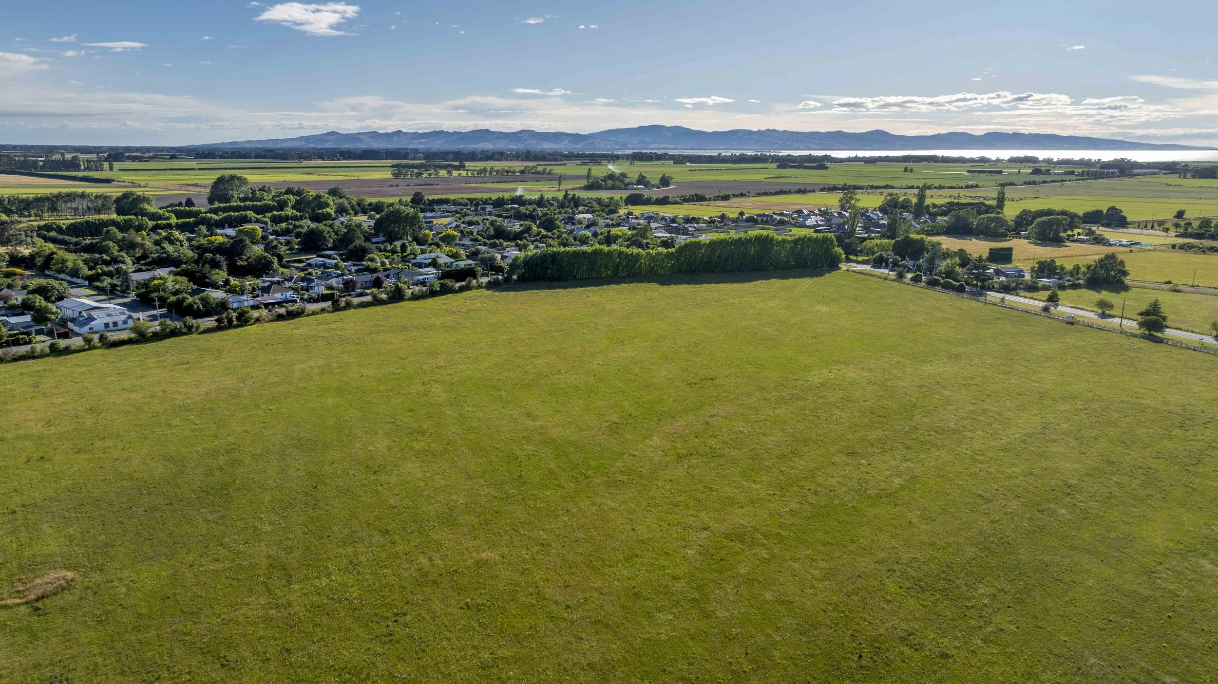 Aerial view of a large green field with a residential area and trees on one side, surrounded by farmland and mountains in the distance. Clear blue sky with scattered clouds.
