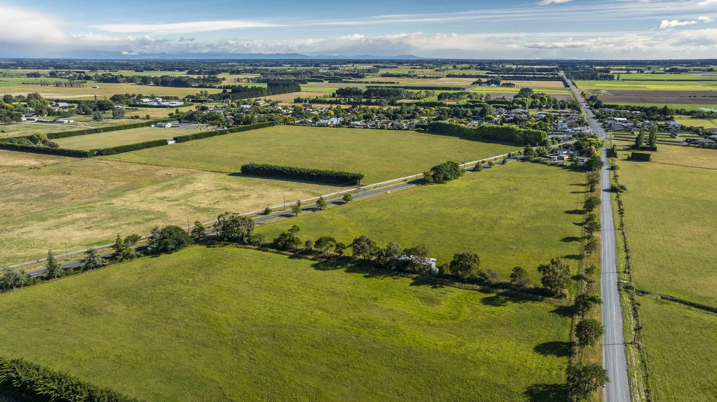 Aerial view of farmland with fields, roads, and a small town in the distance under a partly cloudy sky.