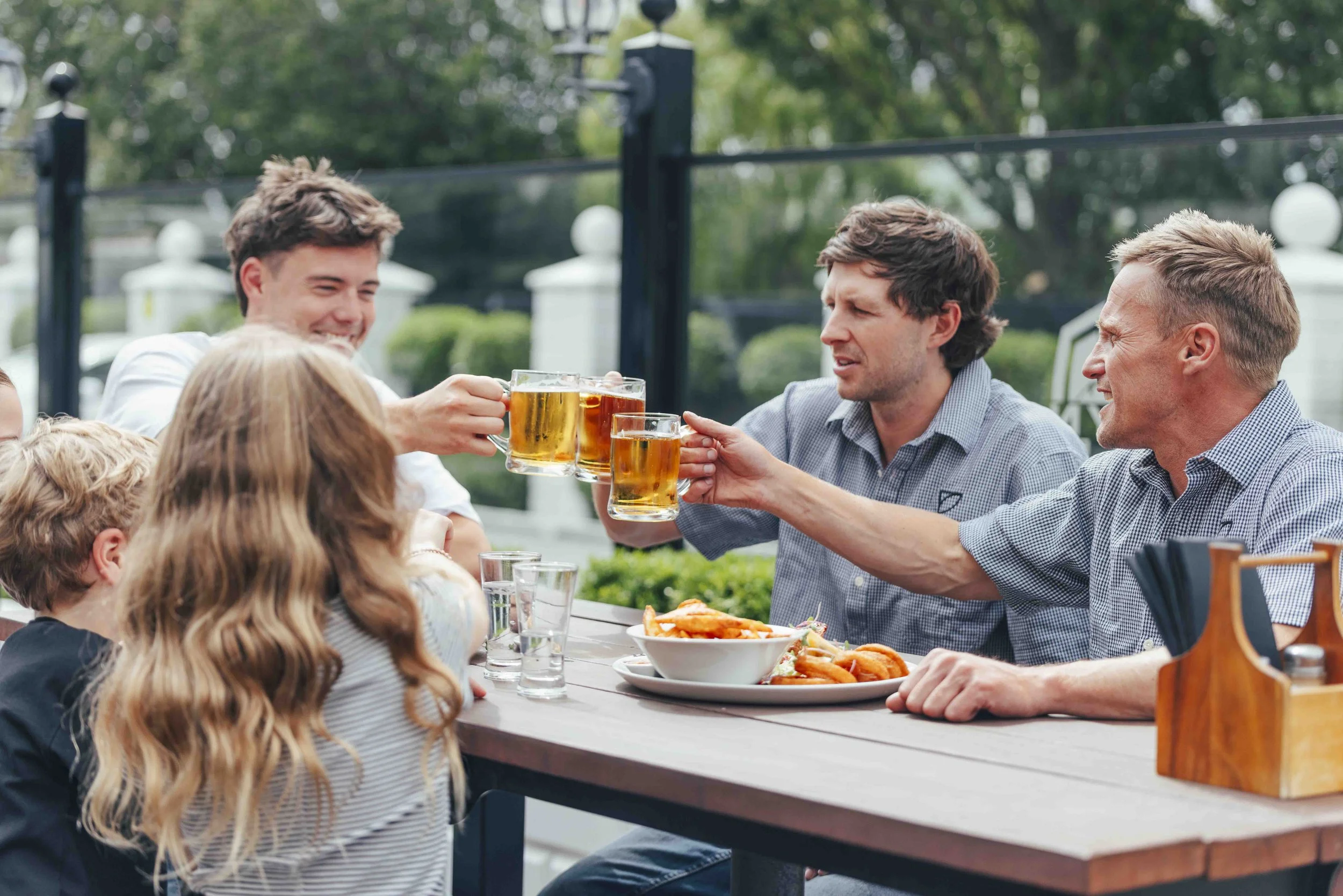 A group of adults and children sitting at an outdoor table, clinking glasses of beer in a toast, with food on the table and a fence with trees in the background.