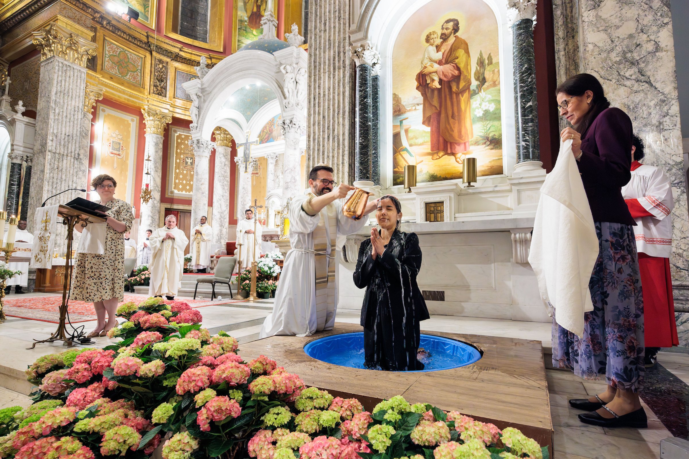 A person in a black dress is being baptized in a church by a man holding a jar of water, with another woman assisting. The church interior features ornate architecture, religious paintings, and floral decorations.