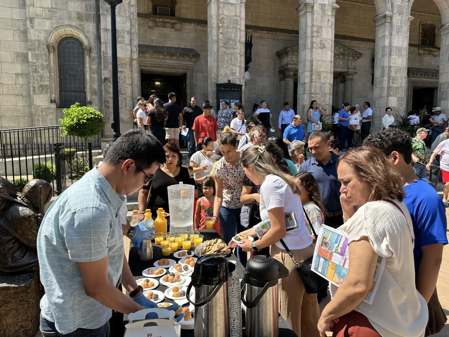People gather around a table with food and drinks outside a historic building in bright sunlight.