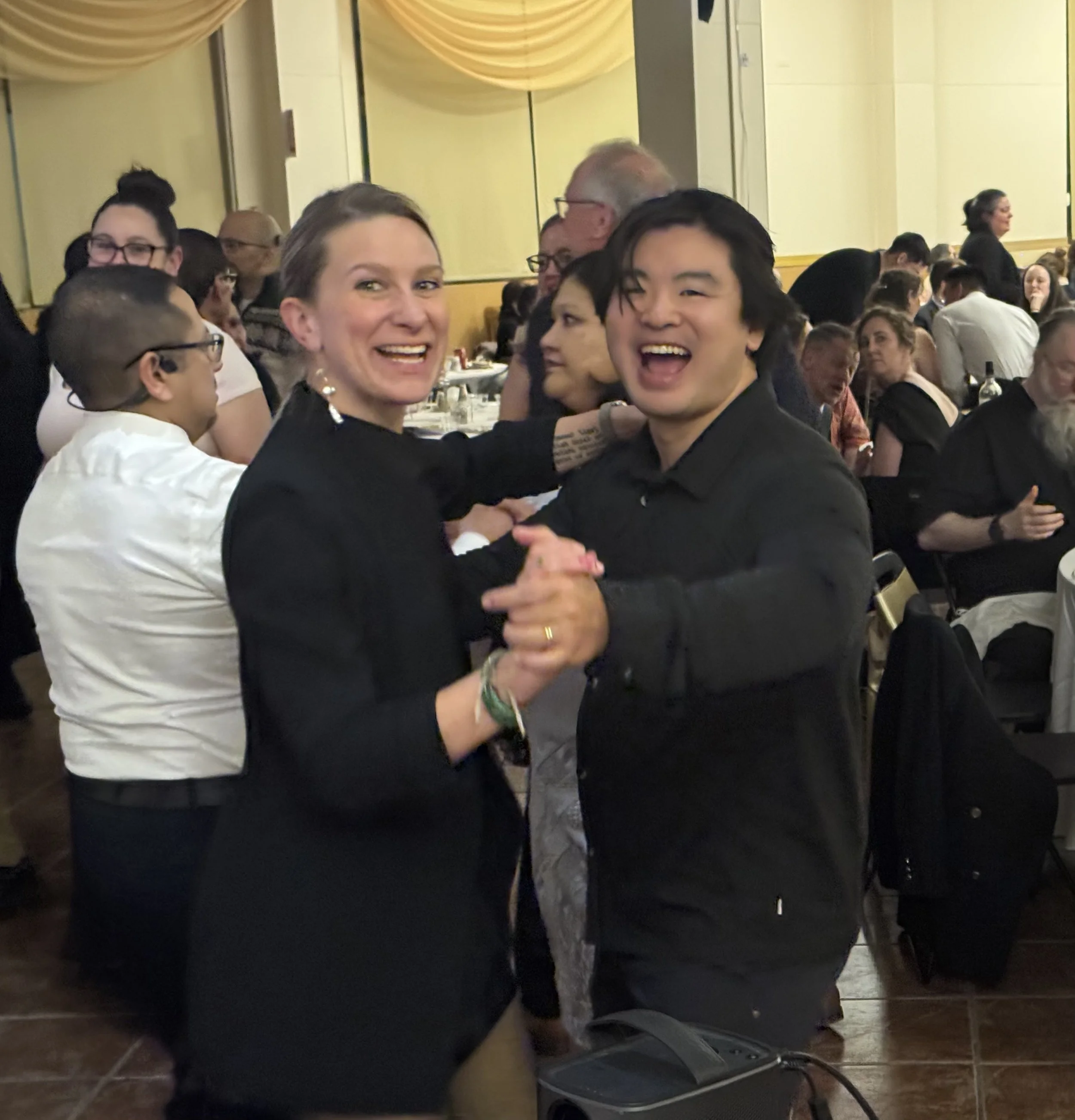 People smiling, greeting, and socializing at a large gathering or event, possibly a banquet or conference, in a decorated hall with yellow drapes.