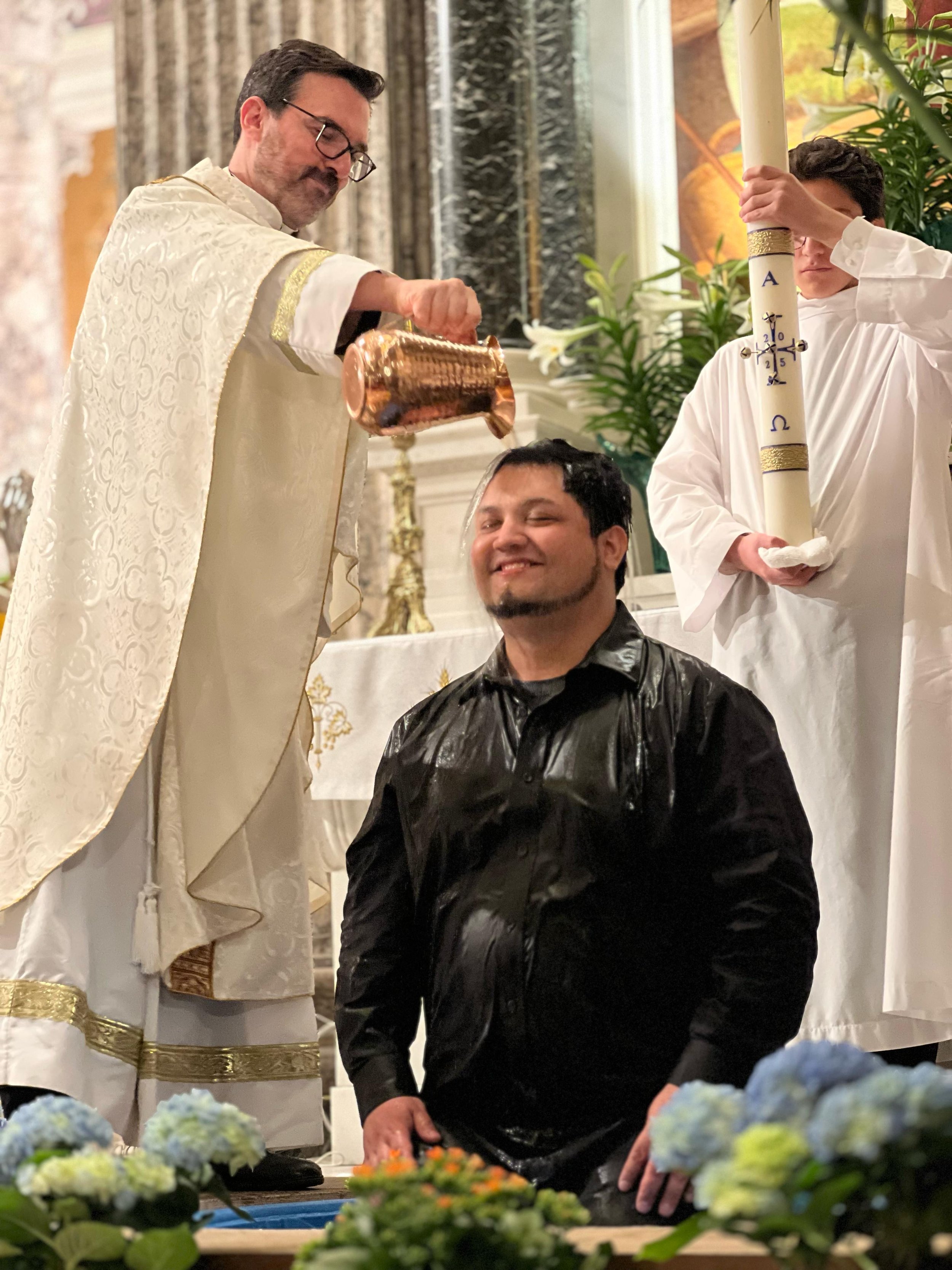 A priest pours water over a man's head during a baptism ceremony in a church with floral decorations and two altar servers present.