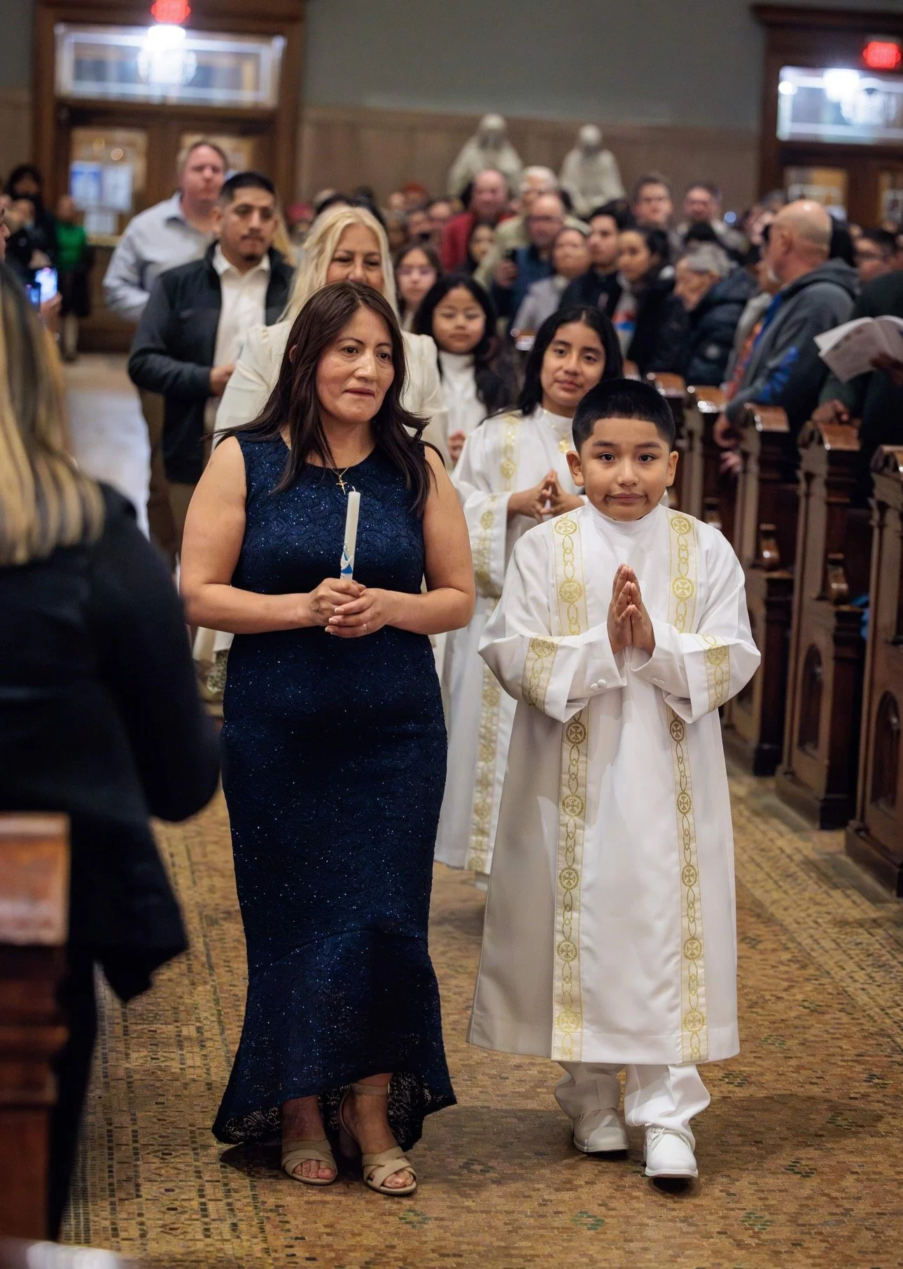 A woman in a dark blue, sparkly evening gown and beige high-heeled sandals walking down a church aisle, holding a candle, followed by a young boy in white religious robes with gold embroidery, in a crowded church with people seated and standing on both sides.
