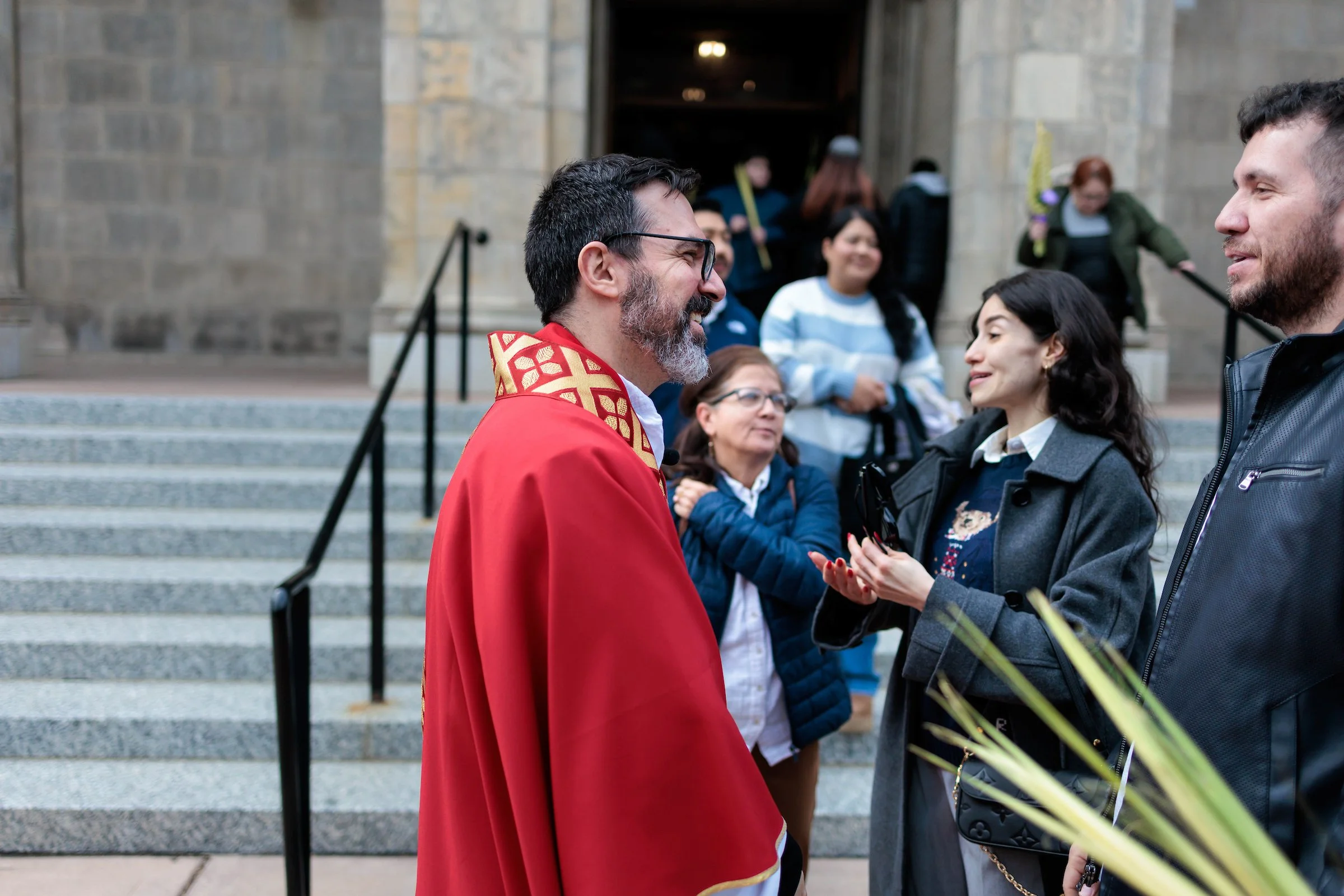 Two men are greeting each other outside a building with steps, smiling and shaking hands. One man is wearing a red robe with gold trim, and the other is dressed casually with a beard and short hair.