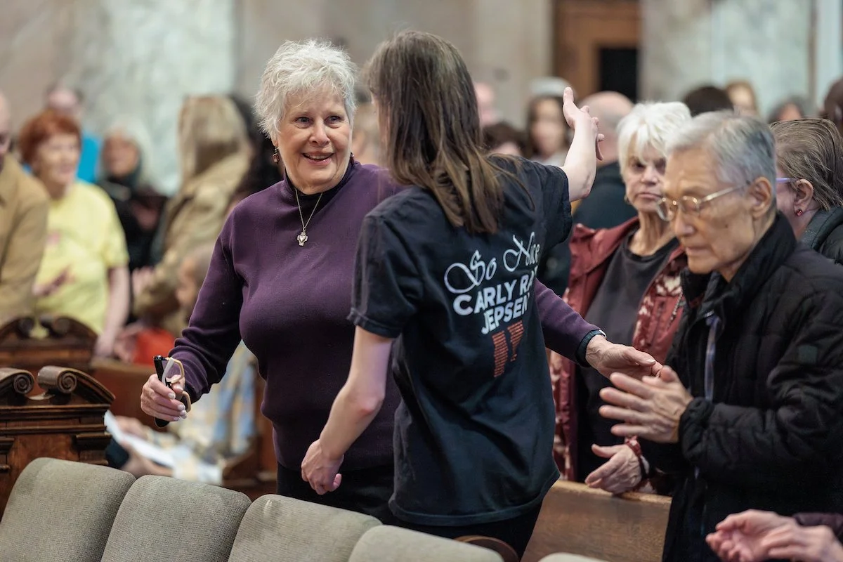 An elderly woman with short gray hair wearing a purple sweater smiling and holding a pen, engaging in conversation with a young woman with long brown hair wearing a black t-shirt with white writing, in a gathering of people in a large hall.