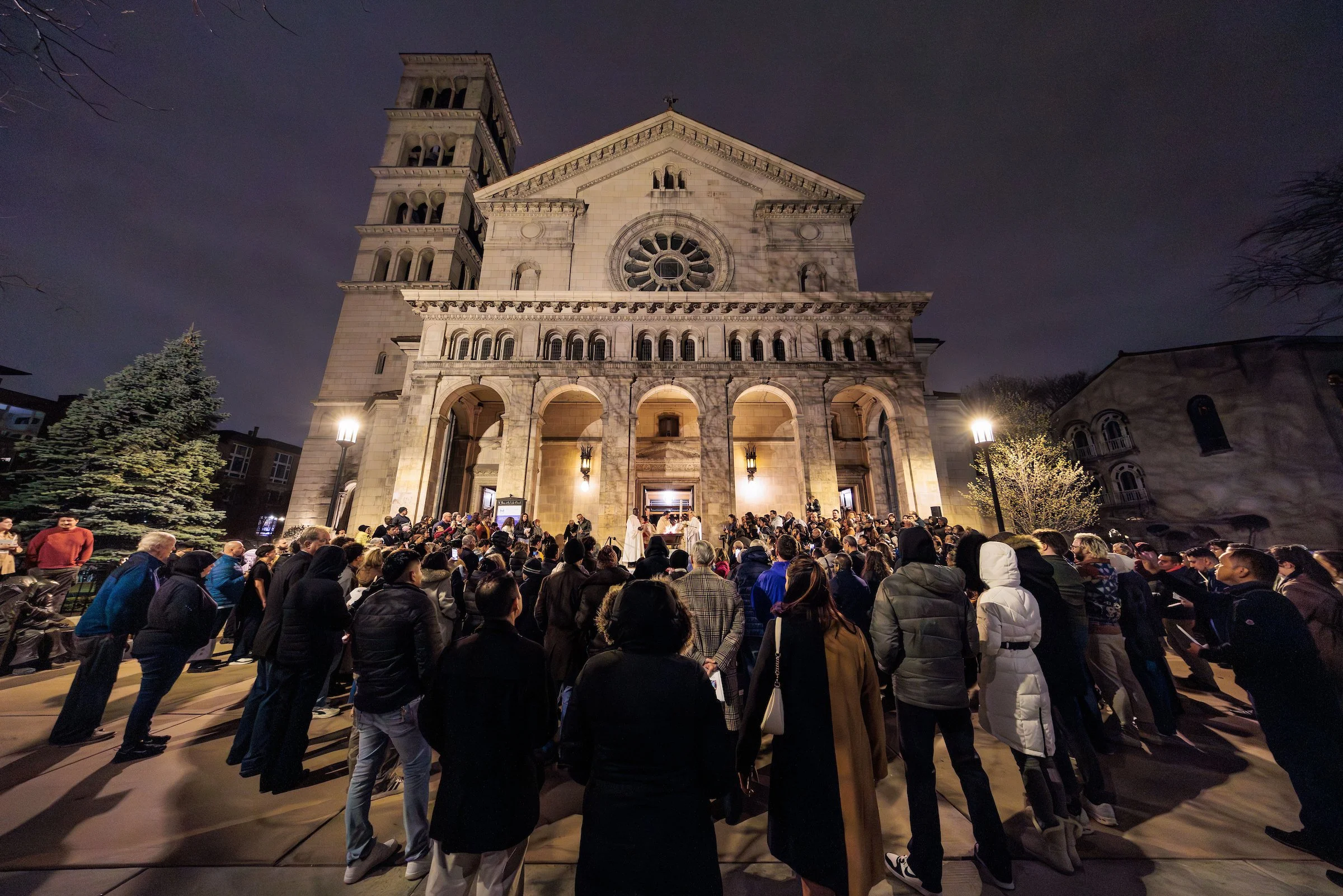 Nighttime scene of a large crowd gathered outside a historic church building with a prominent bell tower and detailed architecture. Some people are seated, and there is a priest or speaker at the front. The church is illuminated, casting shadows on the facade, and trees are visible on the sides. The sky is dark and cloudy.