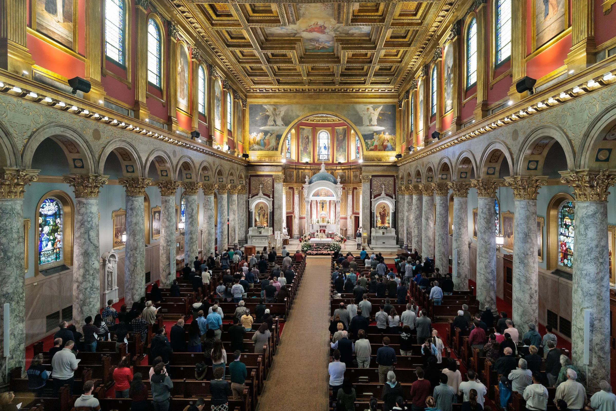 Interior of a church with a high decorated ceiling, large stained glass windows, ornate gold and marble columns, and a congregation gathered facing the altar.