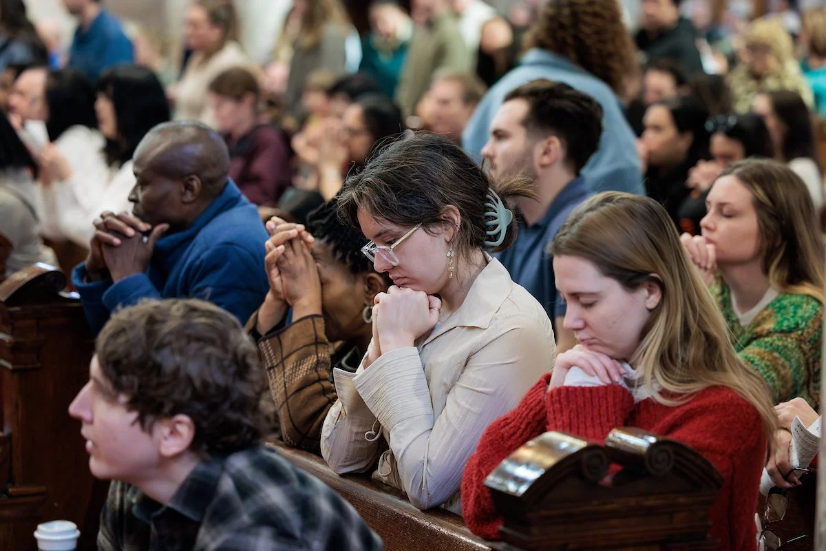 A diverse group of people praying or meditating with eyes closed and hands clasped inside a church.