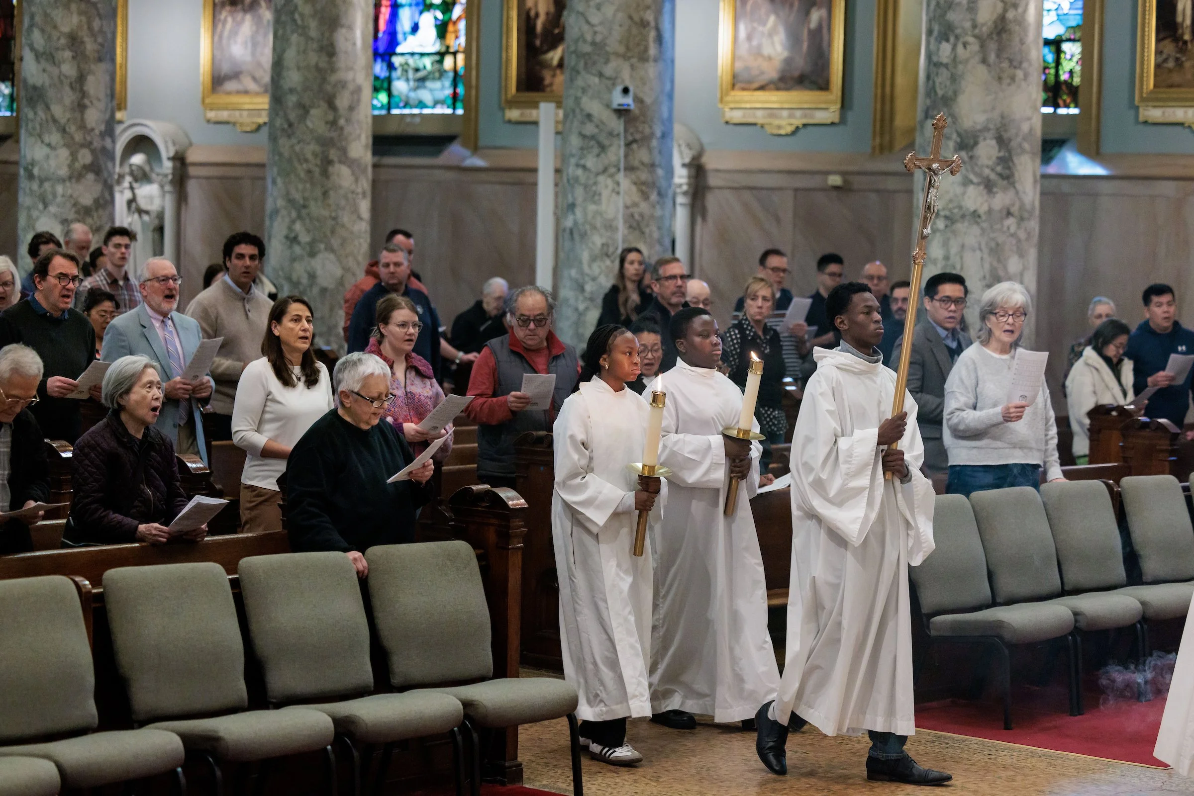 People attending a religious service inside a church, with some standing and singing, including altar servers carrying candles and a crucifix. The church features marble columns, stained glass windows, and framed artwork.