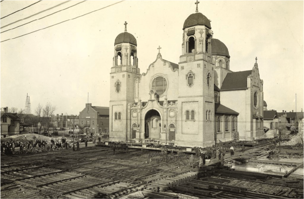 Black and white photo of a church with multiple towers, with railroad tracks and construction in the foreground and a crowd of people nearby.