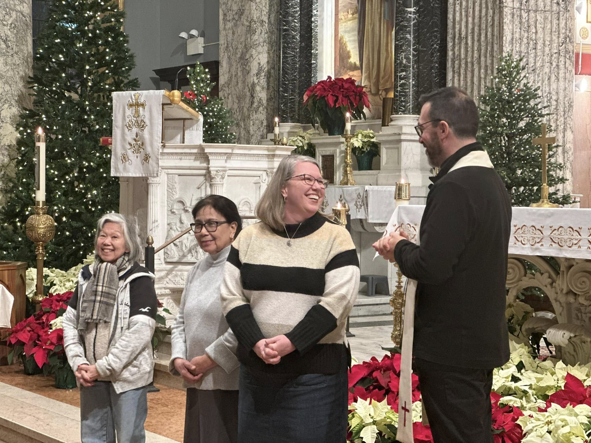 People gathered inside a church decorated for Christmas, with Christmas trees, poinsettias, and candles. Two women are smiling in the foreground, and a man is speaking to a woman who is holding a piece of paper. The altar is decorated with candles, flowers, and a white cloth with gold embroidery.