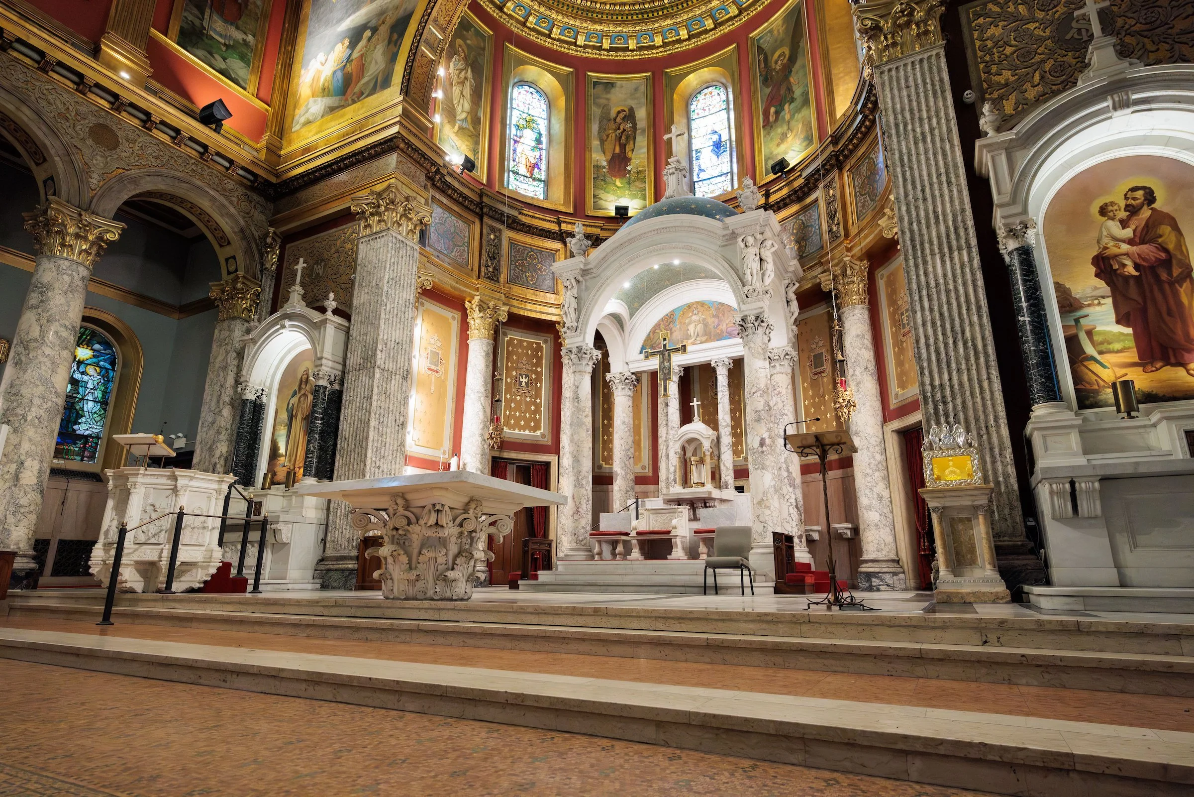 Interior view of a church altar with columns, stained glass windows, and religious artwork on the walls and ceiling.