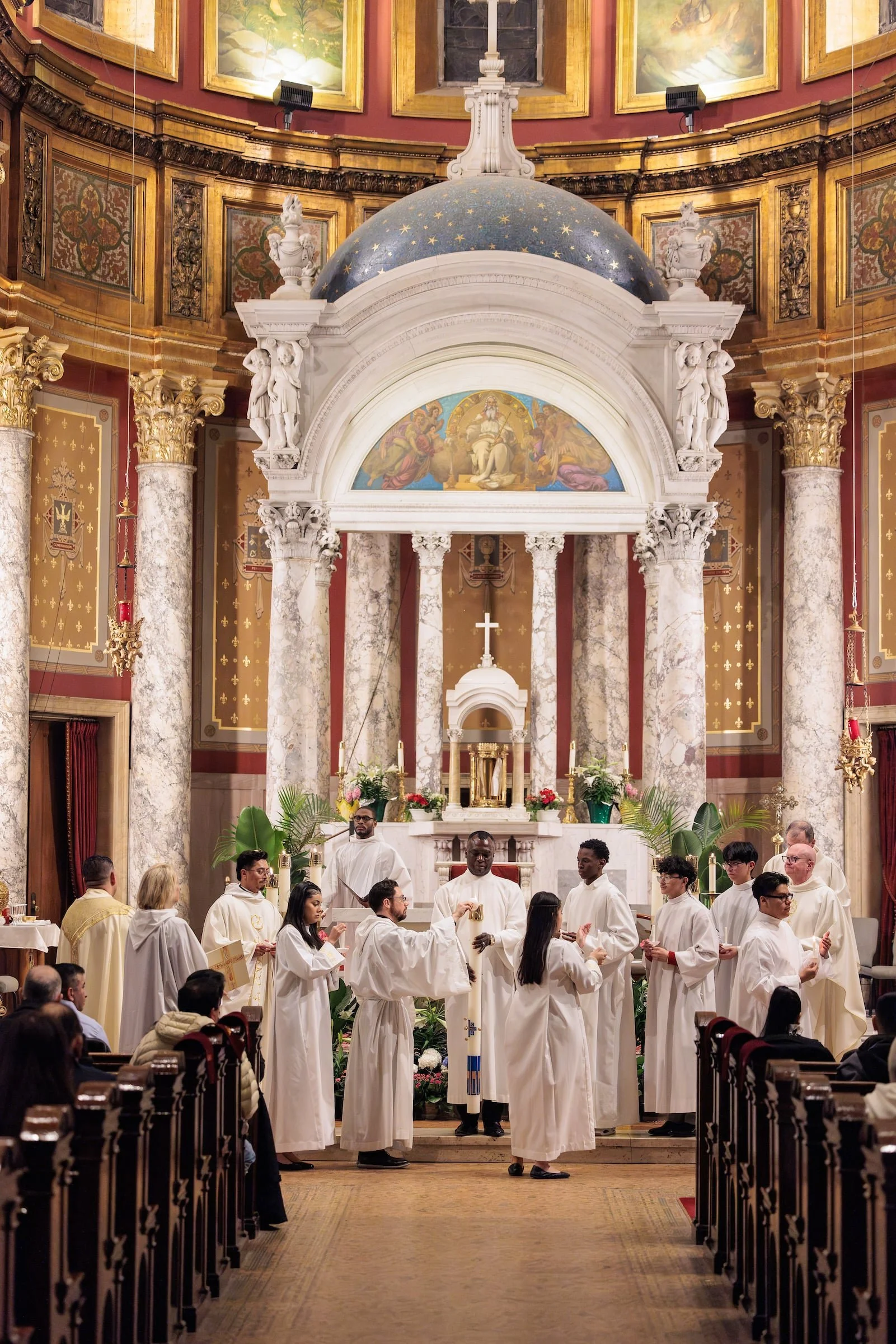 Inside a Catholic church during a religious ceremony with altar servers and clergy members dressed in white robes, standing before the altar adorned with flowers and candles, with ornate gold and marble decorations in the background.