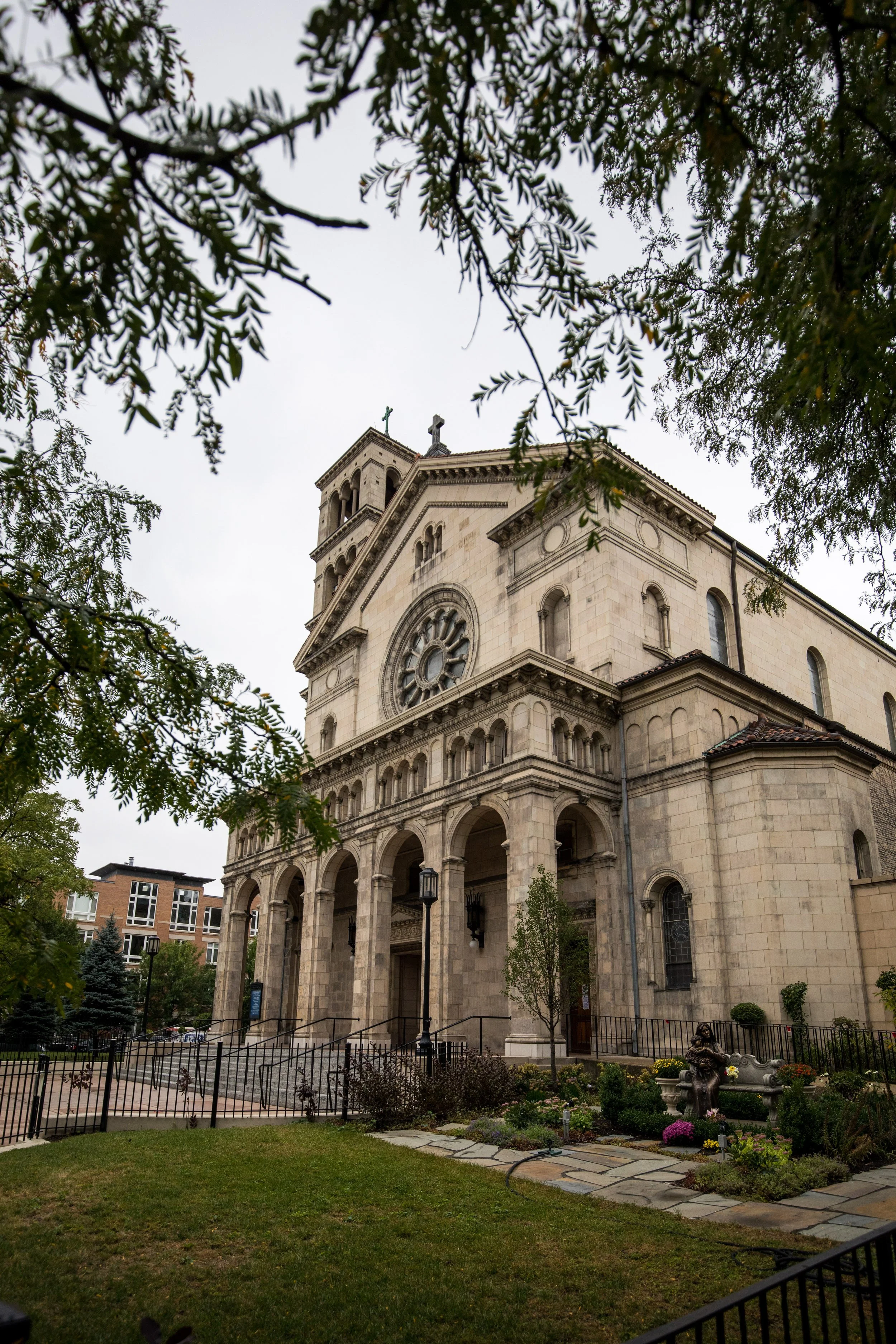 A church building with a tall belfry topped with a cross, arched windows, and a series of large columns at the entrance, seen through trees and greenery in a landscaped area with flowers and sculptures.