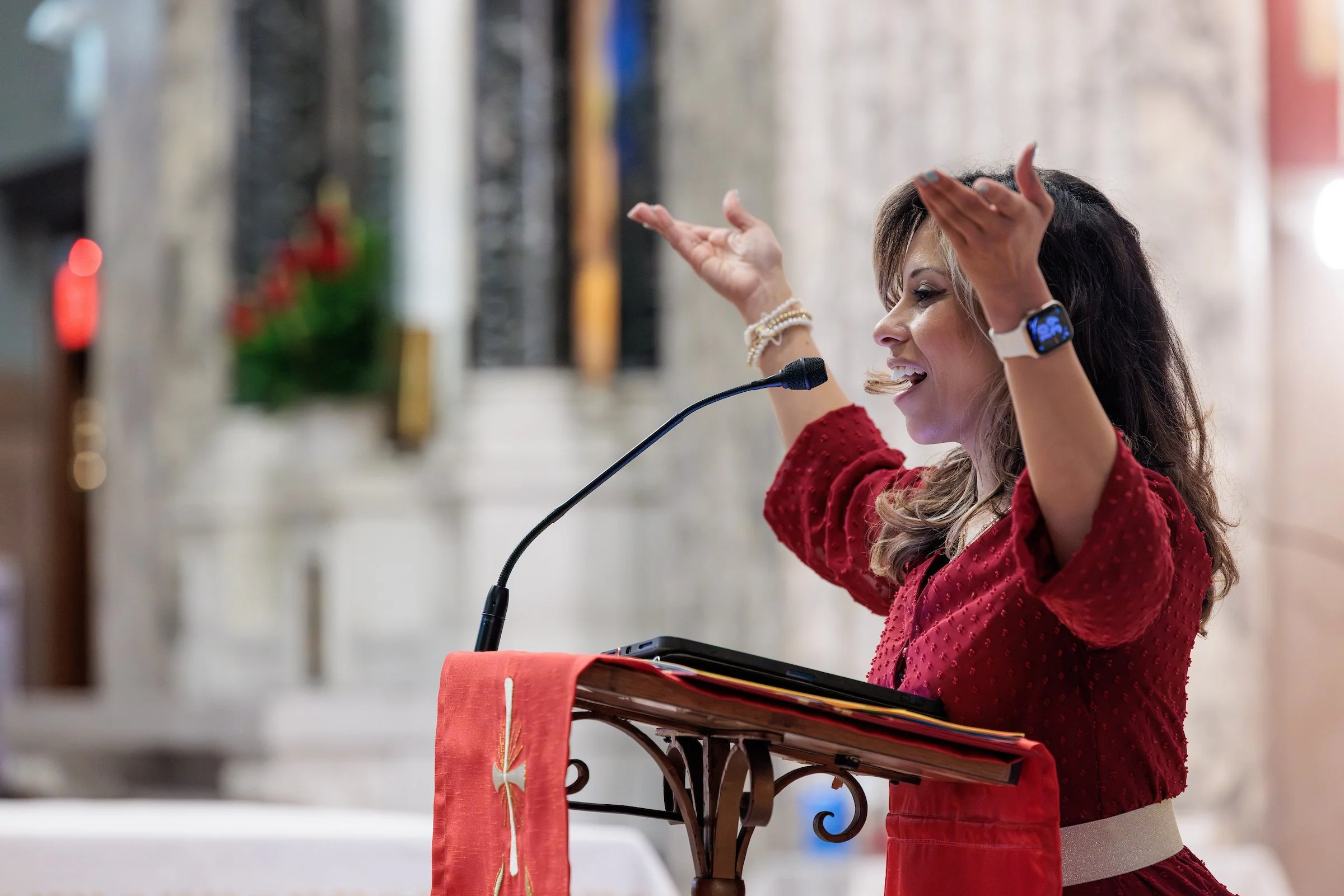 A woman with brown hair and wearing a red dress is speaking at a podium with her hands raised. She appears to be engaged and expressive.