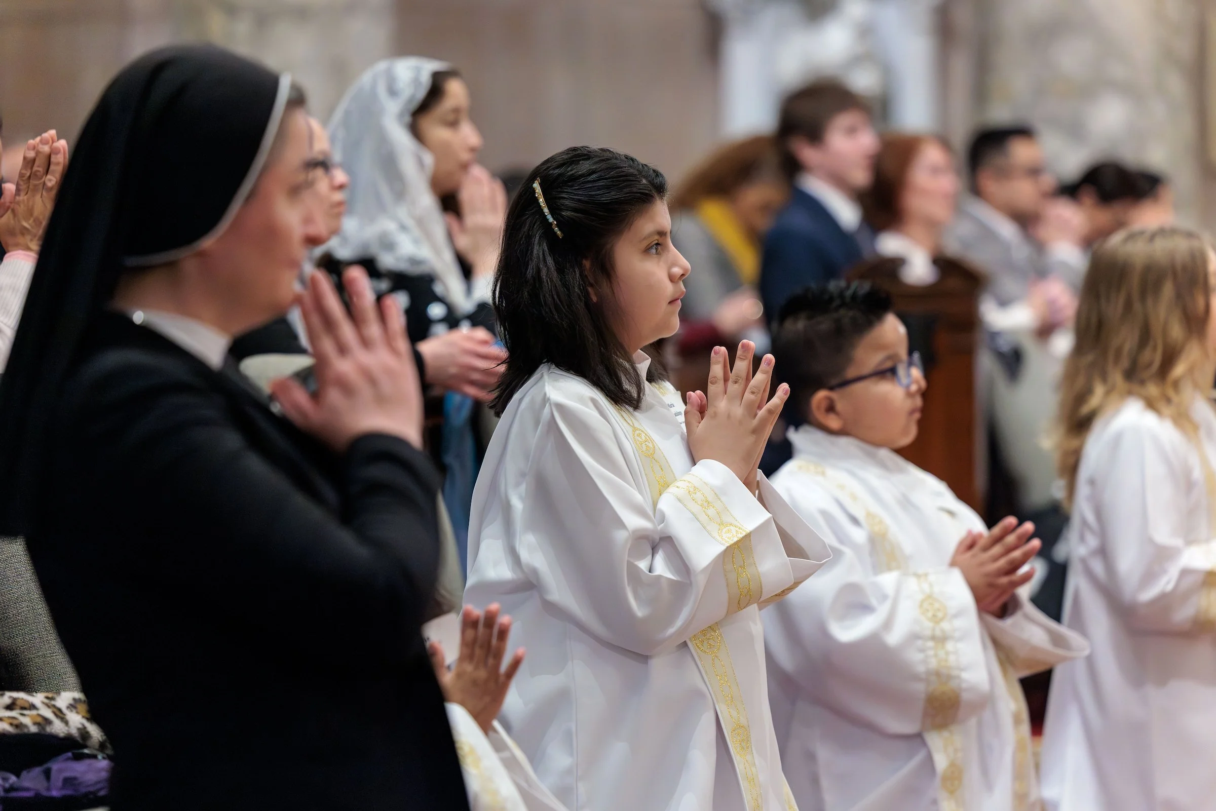 People, including children and women in religious attire, praying with hands clasped during a religious service or ceremony.