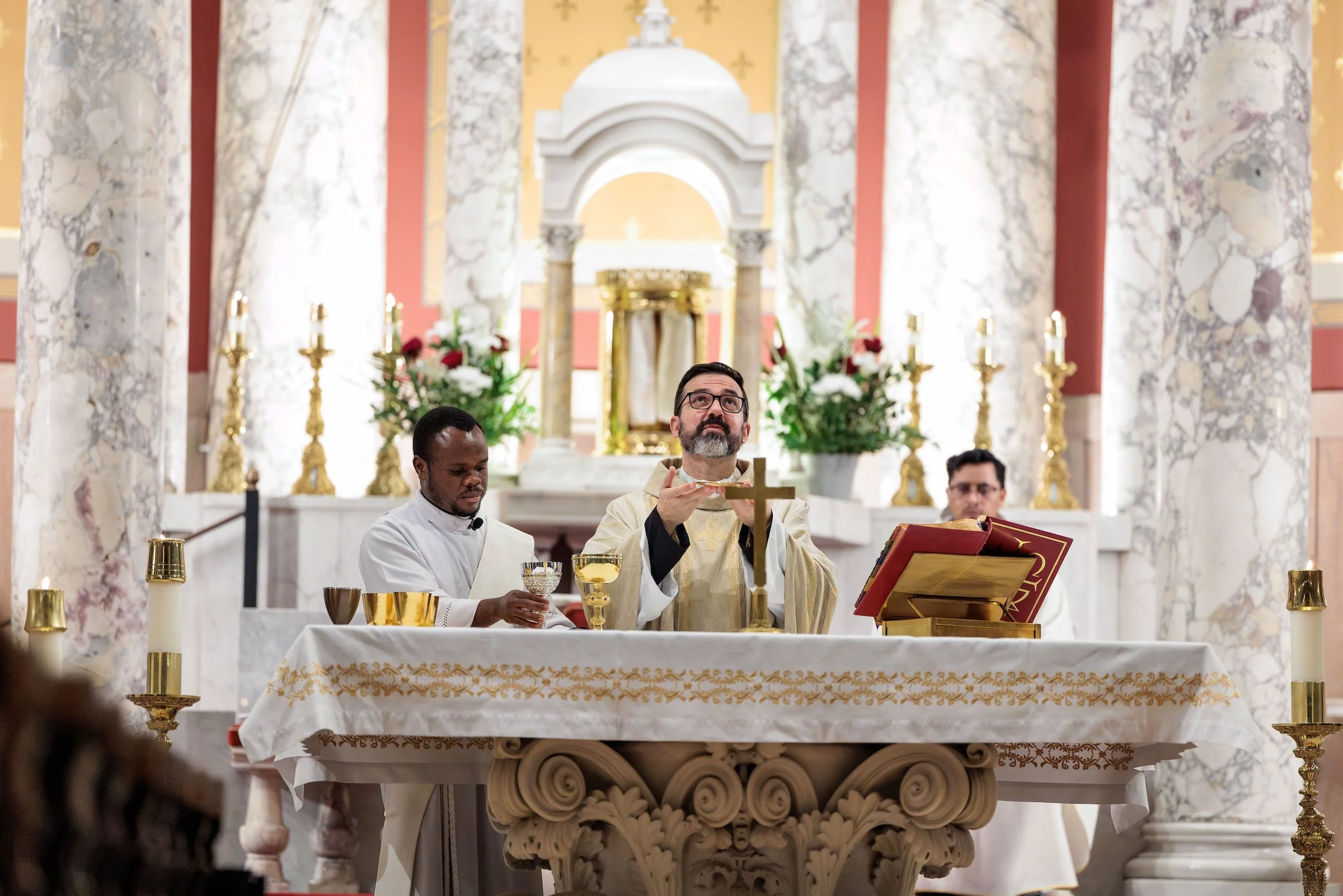 Religious ceremony with a priest and two men sitting at an altar in a church, surrounded by candles and flowers.