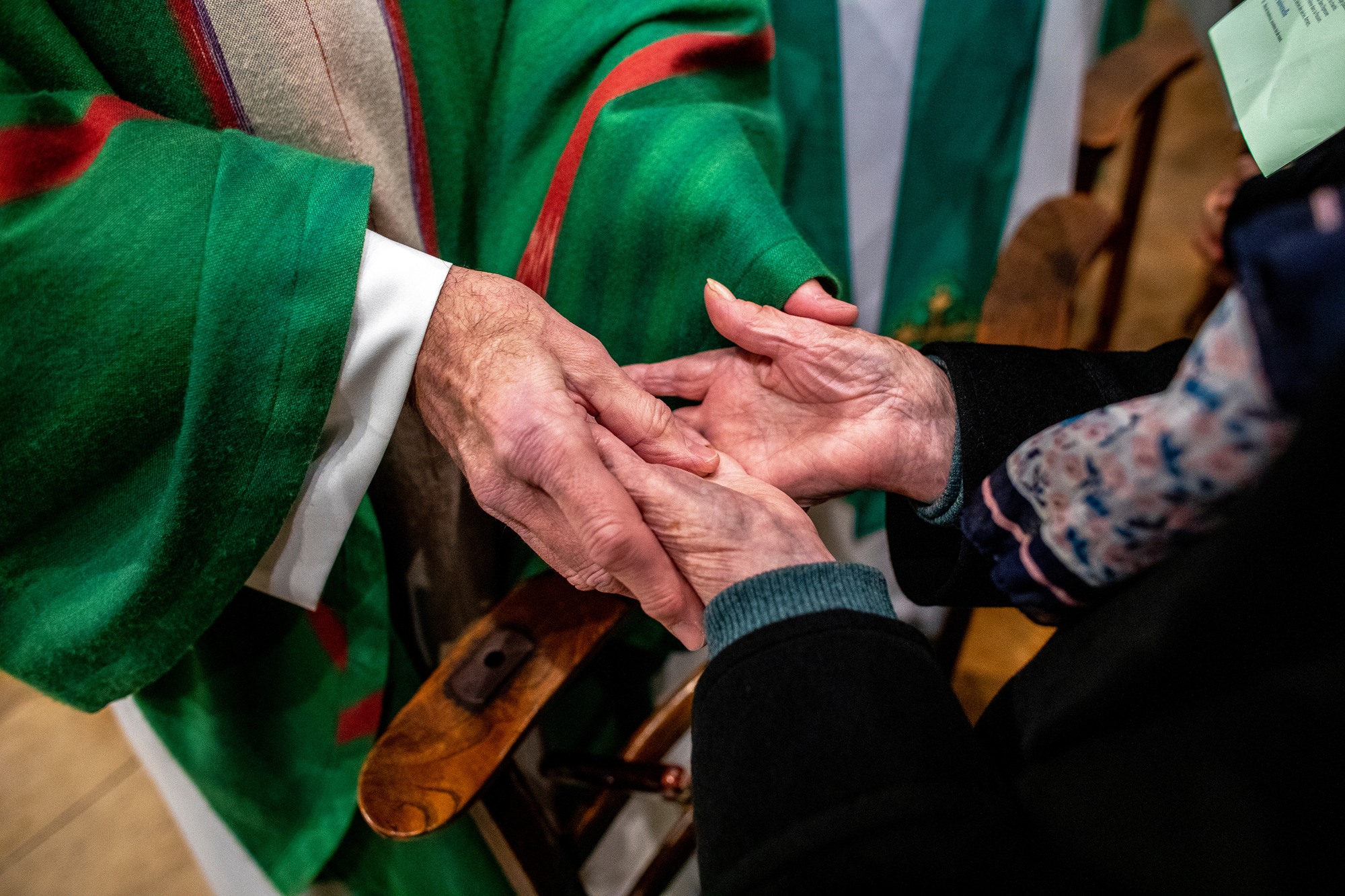 Two people, likely a man and a woman, holding hands in a gesture of greeting or affection, with a woman wearing a festive green and red sweater and part of a wooden bench visible in the background.