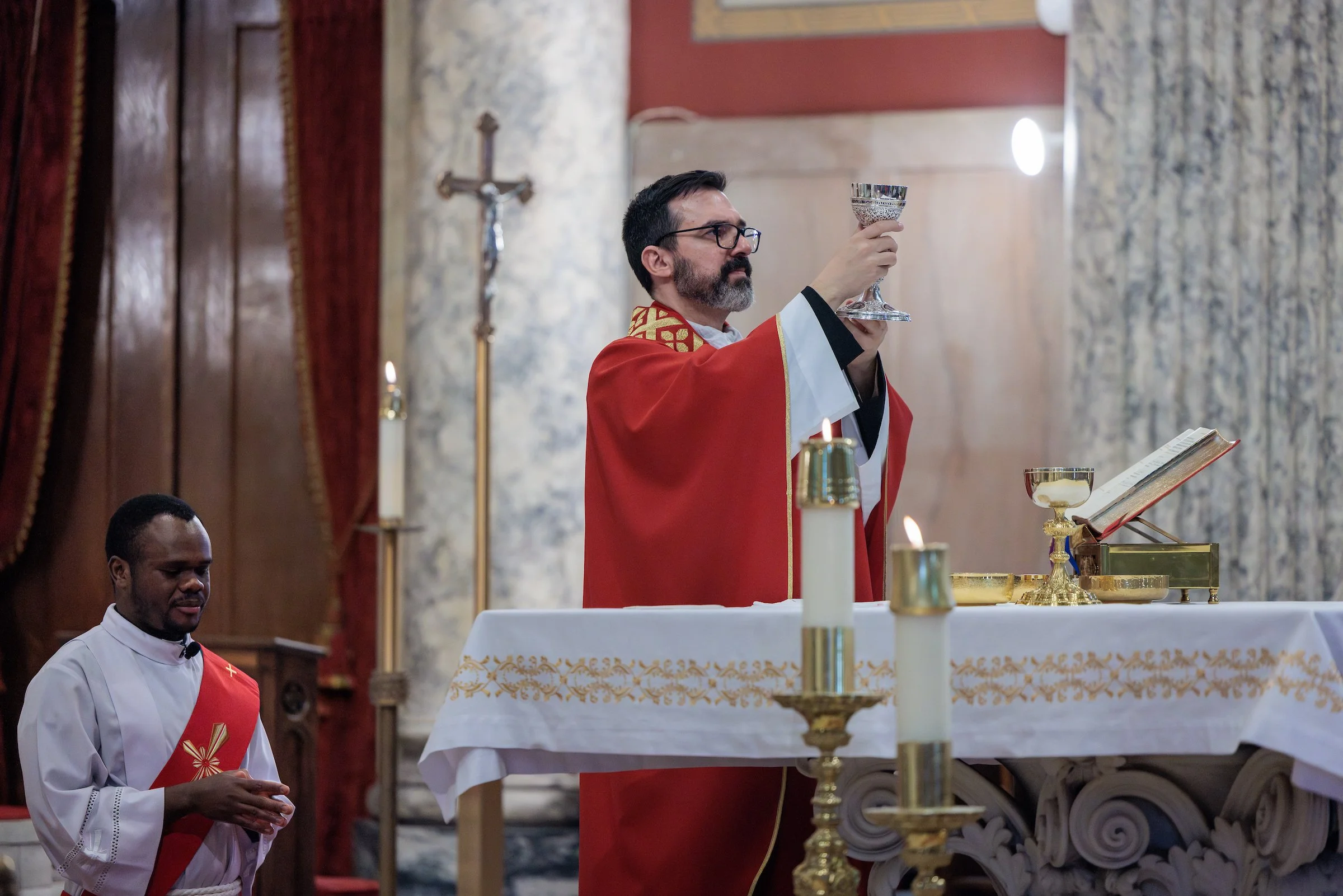 A priest in red vestments holding a chalice during a religious service inside a church, with another altar server standing nearby, and religious items on the altar.