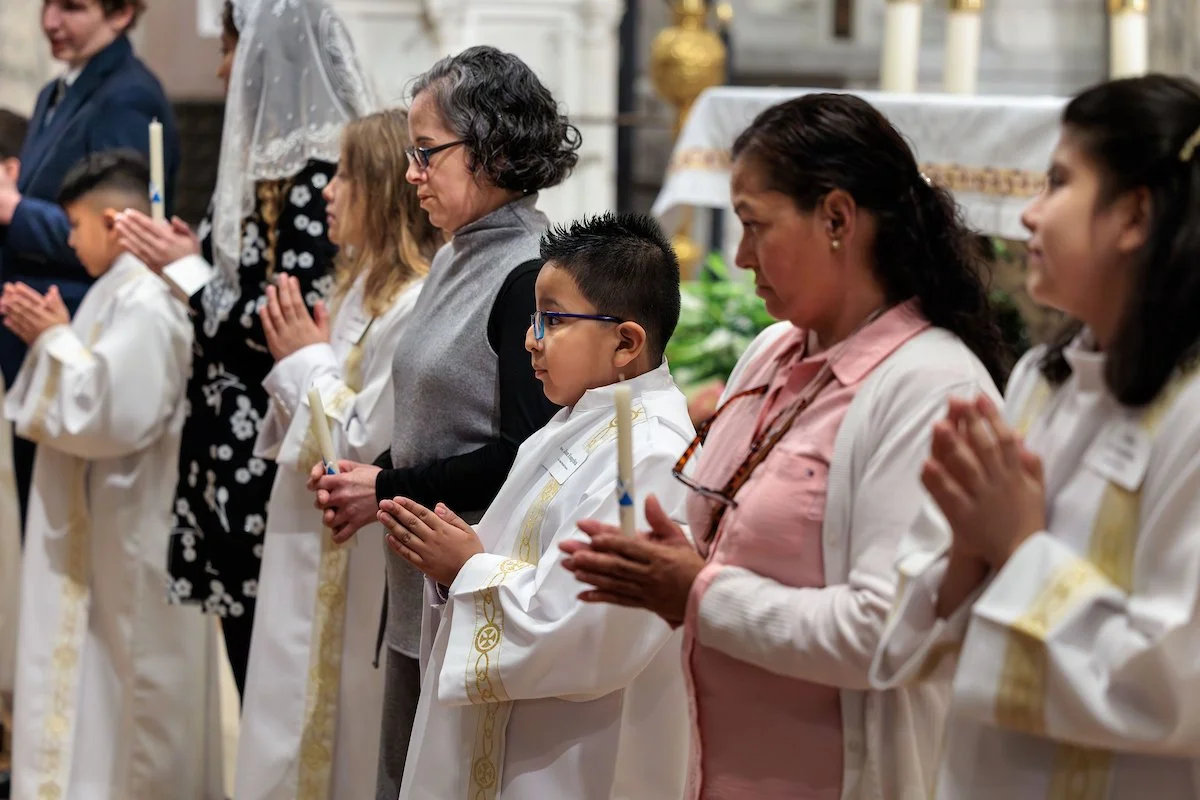 People praying during a religious ceremony in a church, with some holding candles and dressed in white robes.