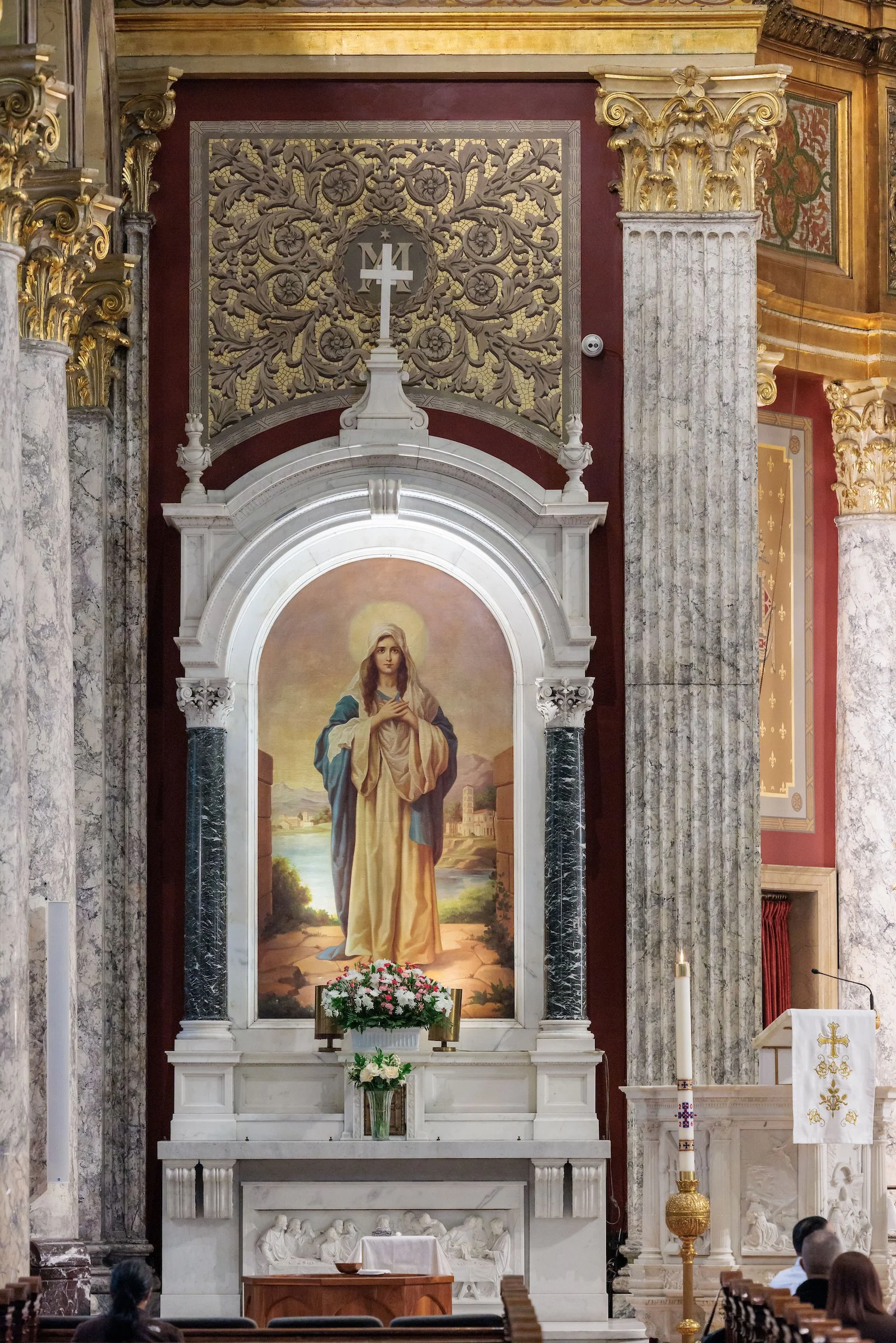 Inside a church, a large painting of Jesus Christ, with a halo, stands surrounded by an ornate altar with columns, flowers, and candles. Religious symbols and intricate gold decorations are visible on the walls.