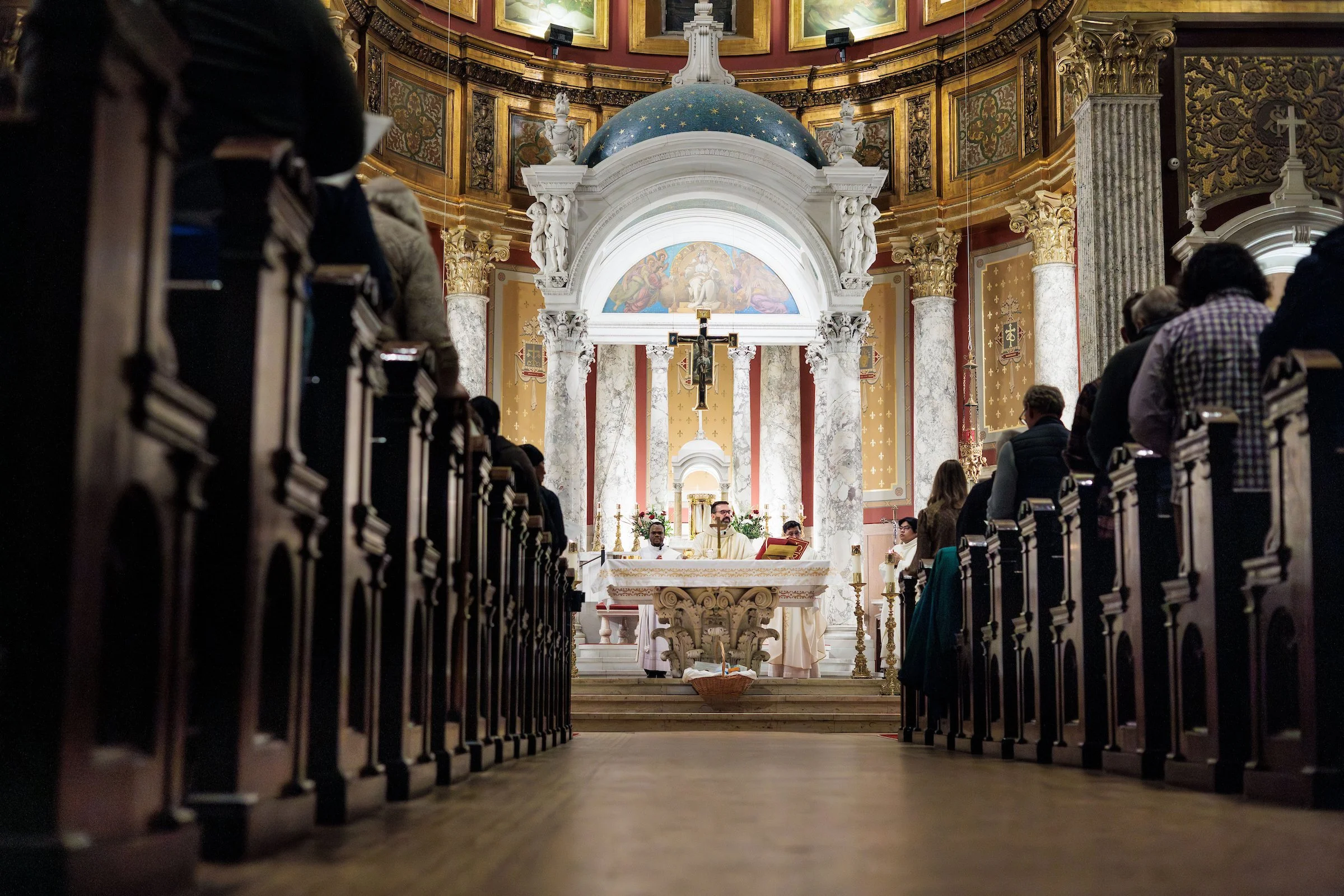 Inside a church with a congregation seated in pews, priests conducting a service at the altar, ornate architecture, columns, religious art, and a crucifix hanging above.