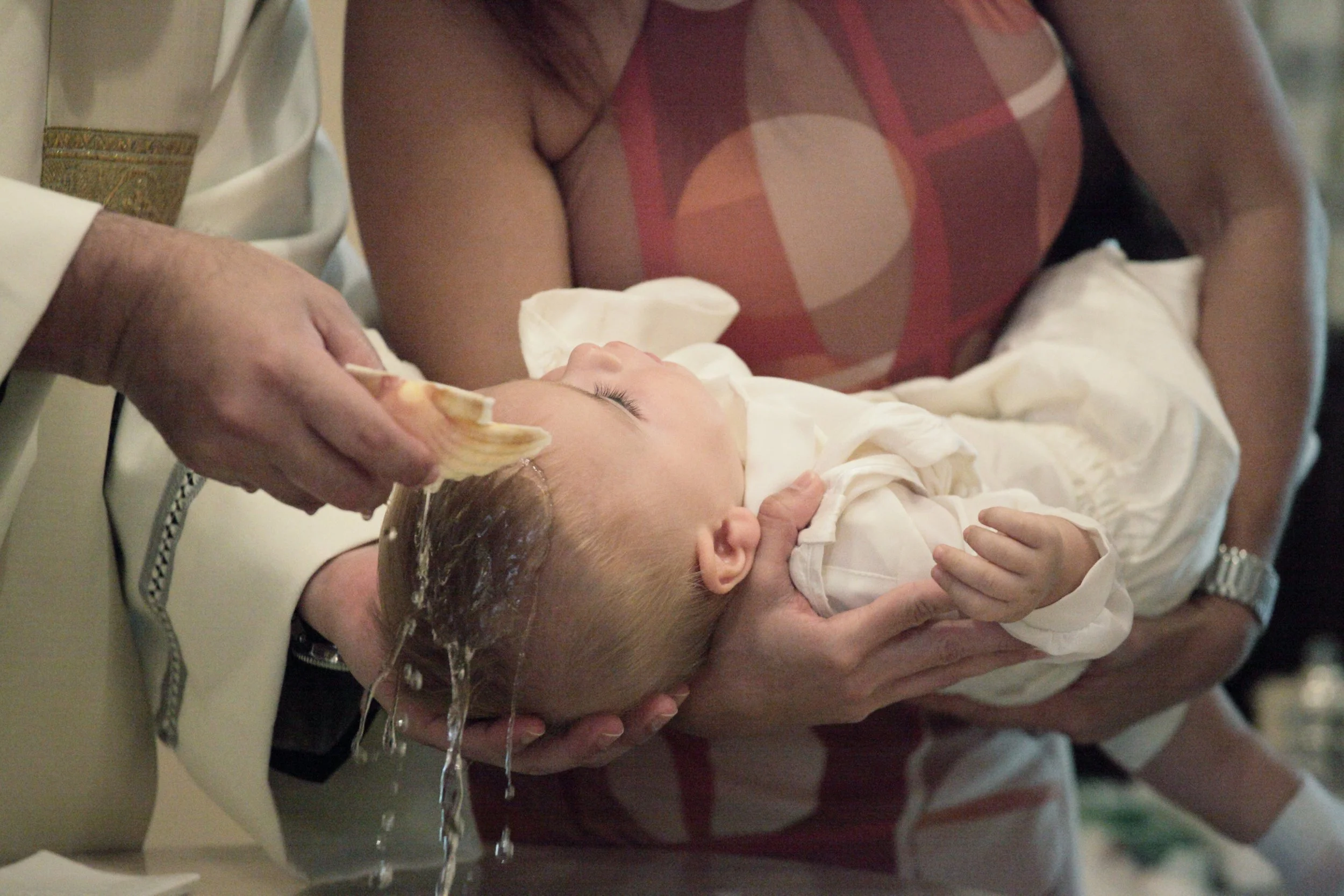 A baby is being bathed, lying in a white basin. An adult's hands are gently washing the baby's head with a sponge or cloth, with water running over the baby's hair. The background includes an adult wearing a sleeveless top with a geometric pattern, and another person in a white shirt.