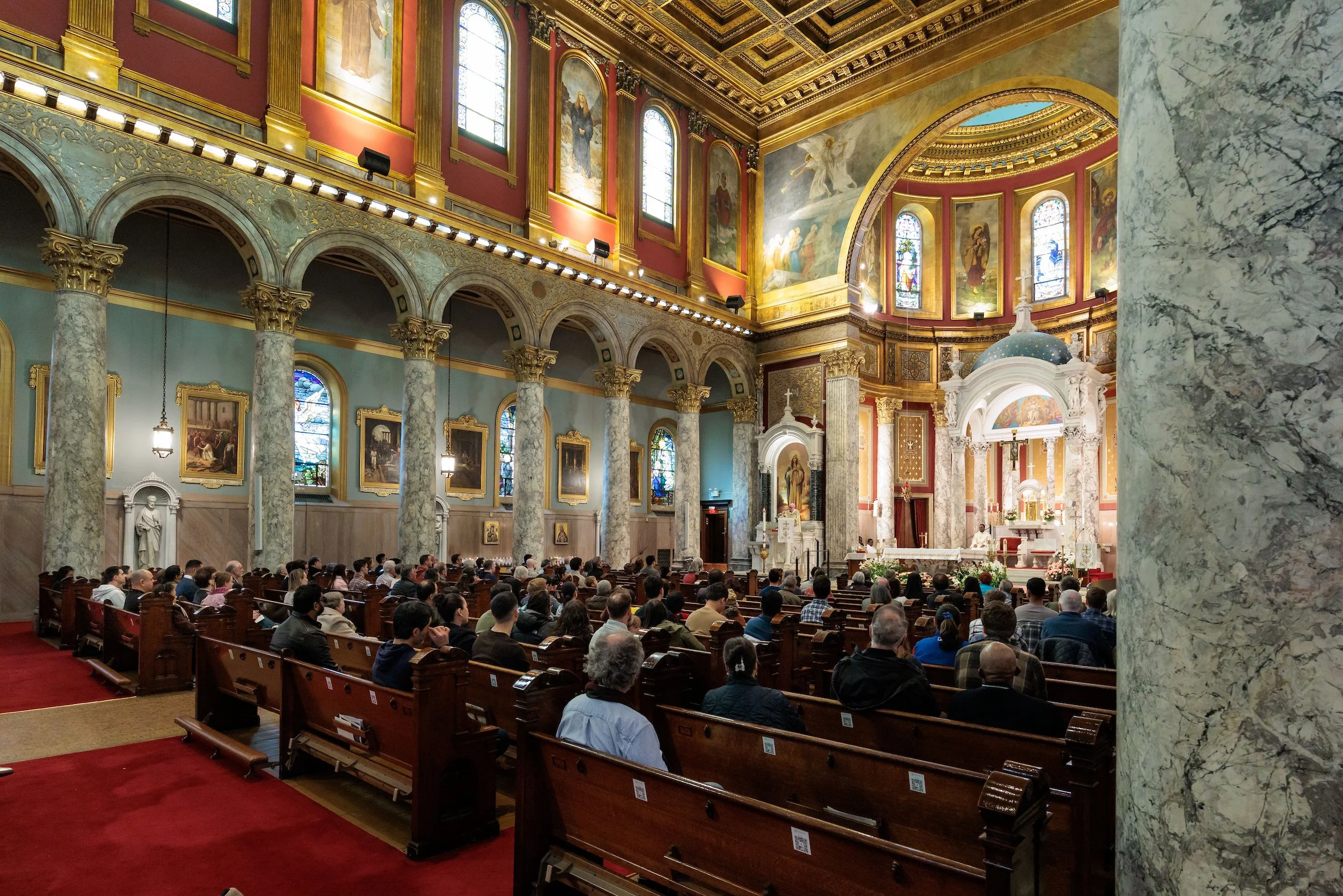Interior of a church with ornate gold and red decorations, stained glass windows, and a congregation seated in pews.