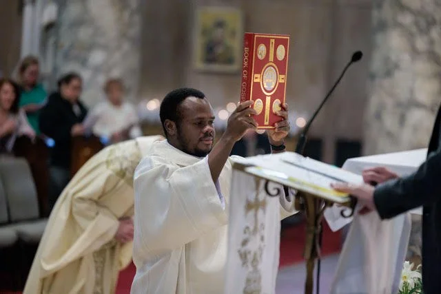 A man dressed in white robes is holding up a Bible with a red and gold cover during a religious ceremony in a church, with others seated in the background.