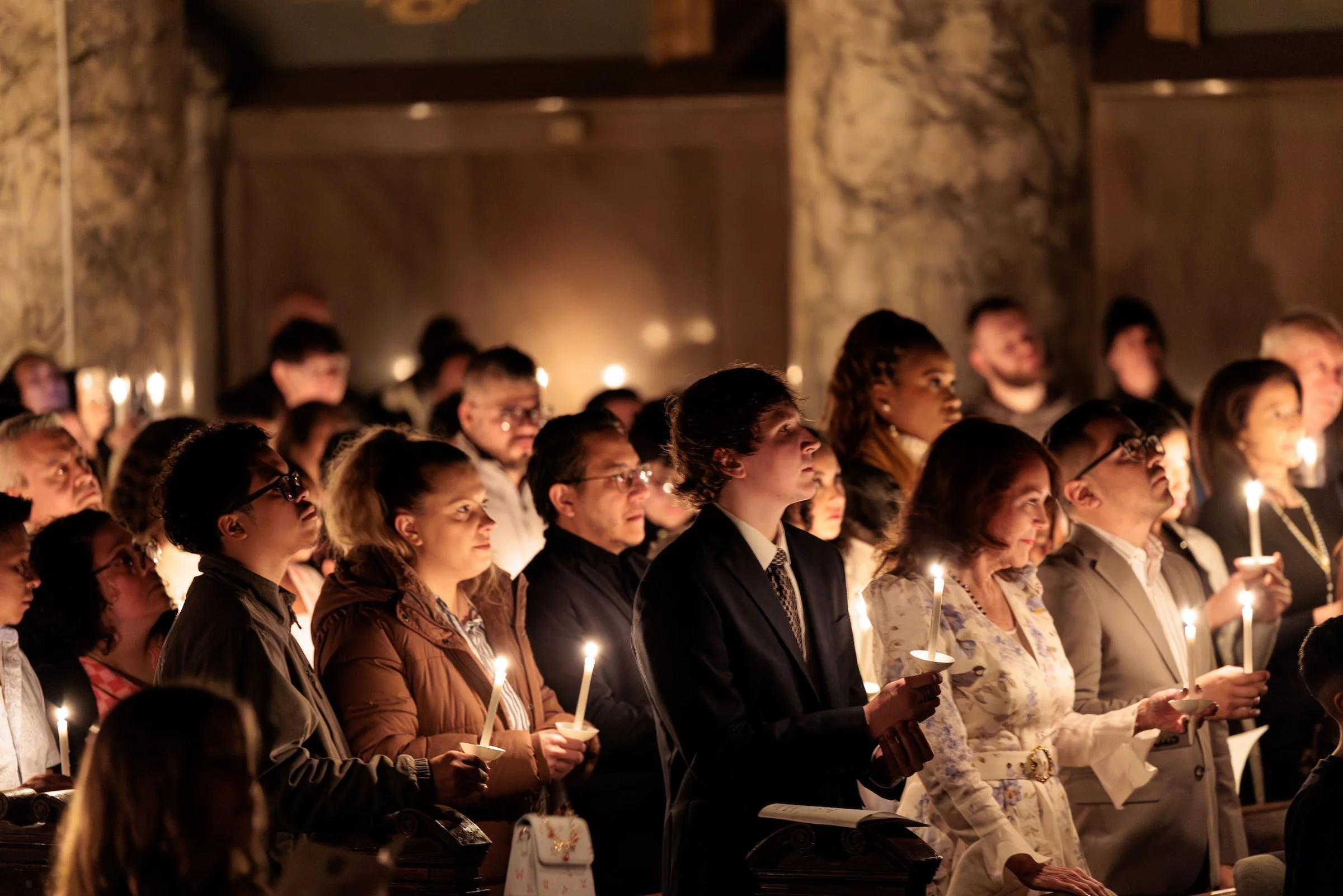 A large group of people attending a candlelight vigil or ceremony, holding lit candles and standing in a dimly lit, ornate room.