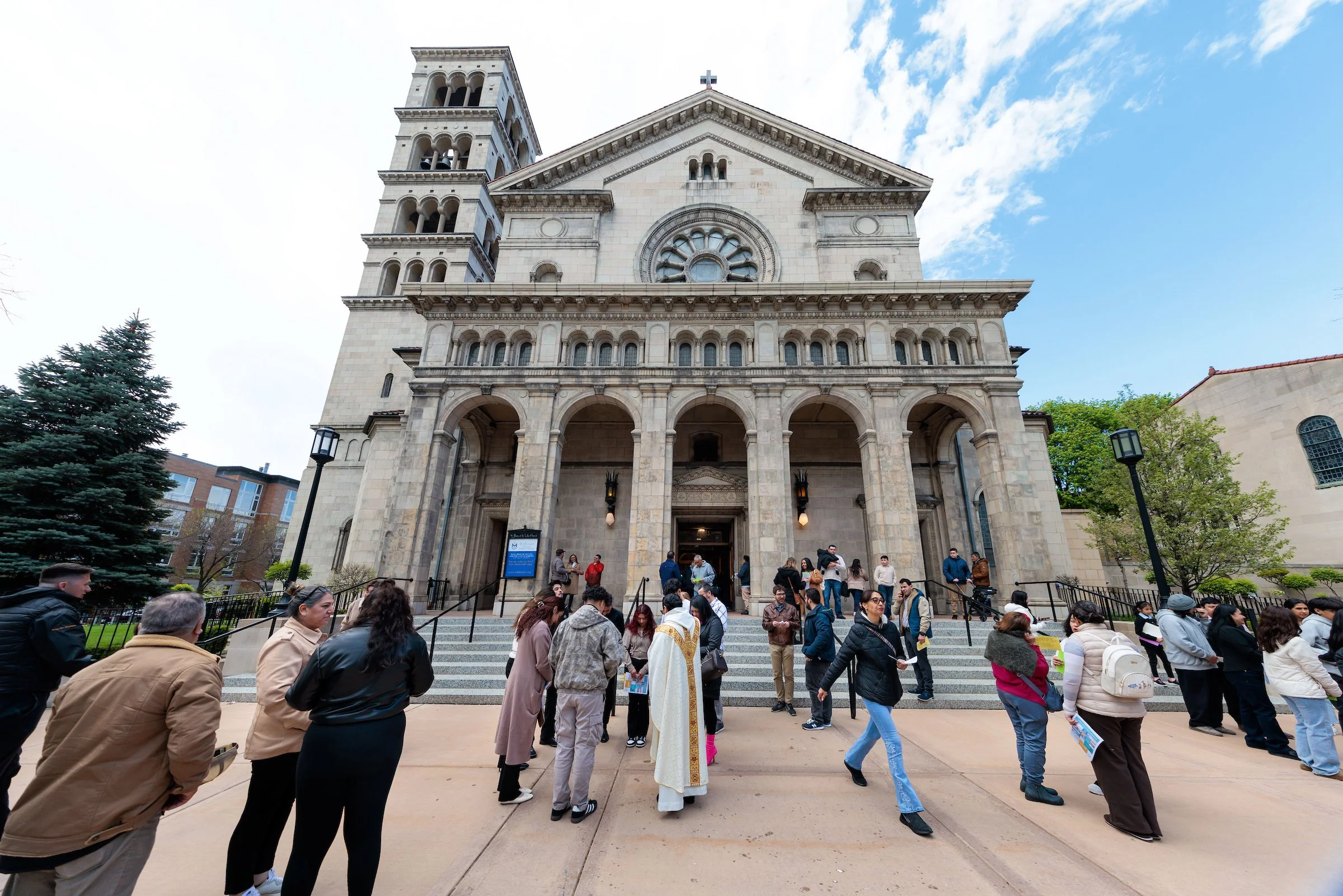 People gathering outside a large stone church with multiple arches and a tall bell tower, under a partly cloudy sky.