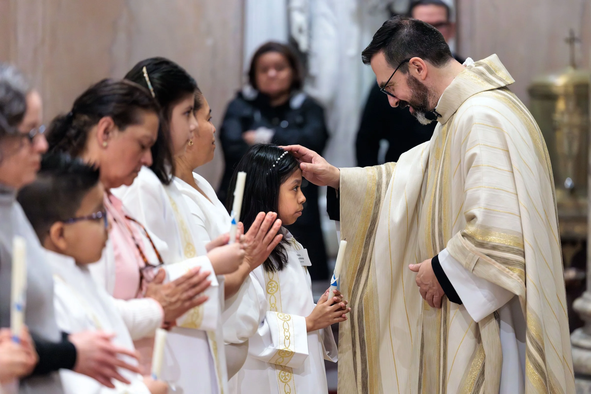 A priest is touching a young girl's forehead during a religious ceremony, with women standing in line in white robes holding candles.