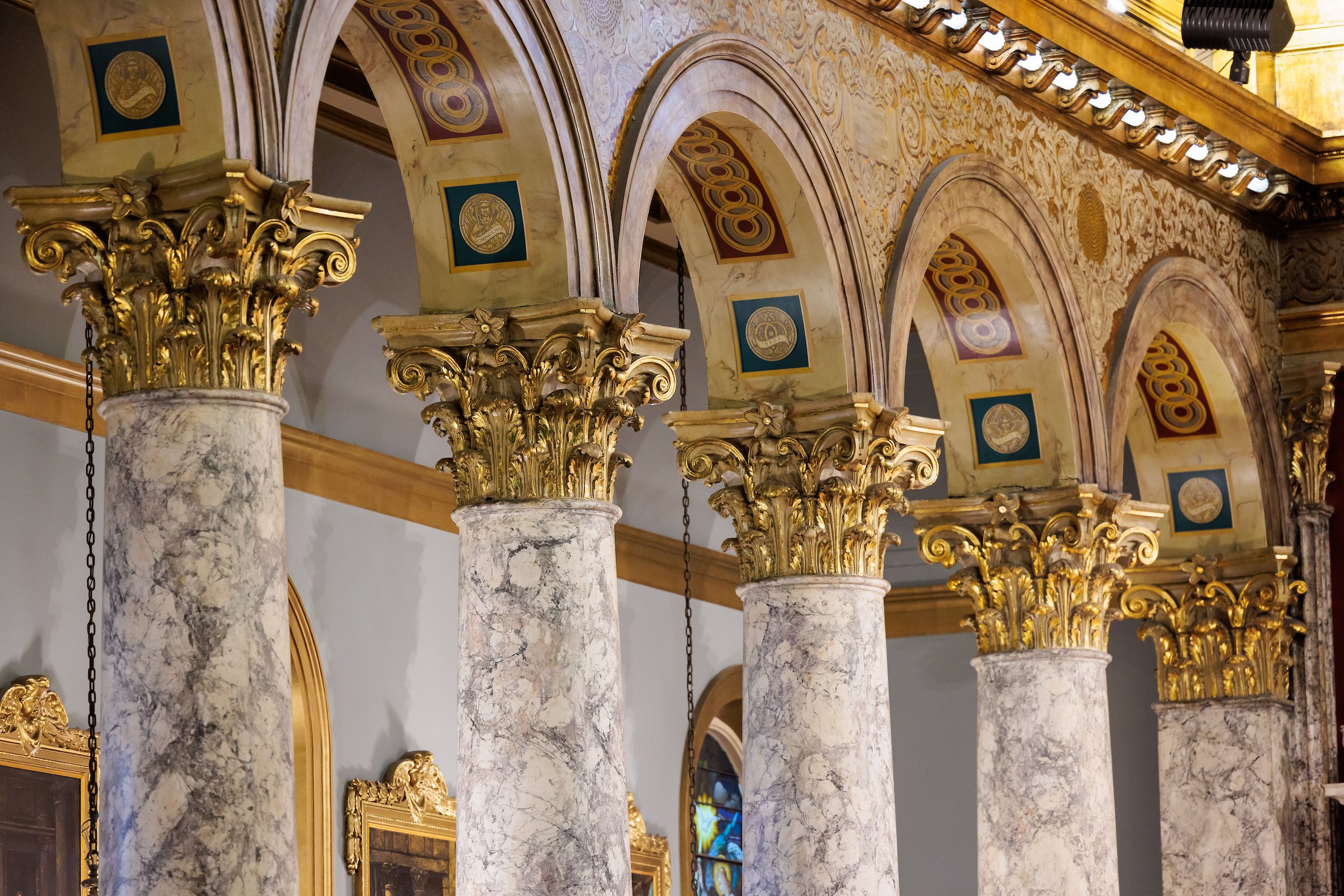 Interior view of a church with ornate gold and marble columns supporting arched ceilings decorated with intricate gold patterns and medallions. The ceiling is illuminated by a row of spotlights.