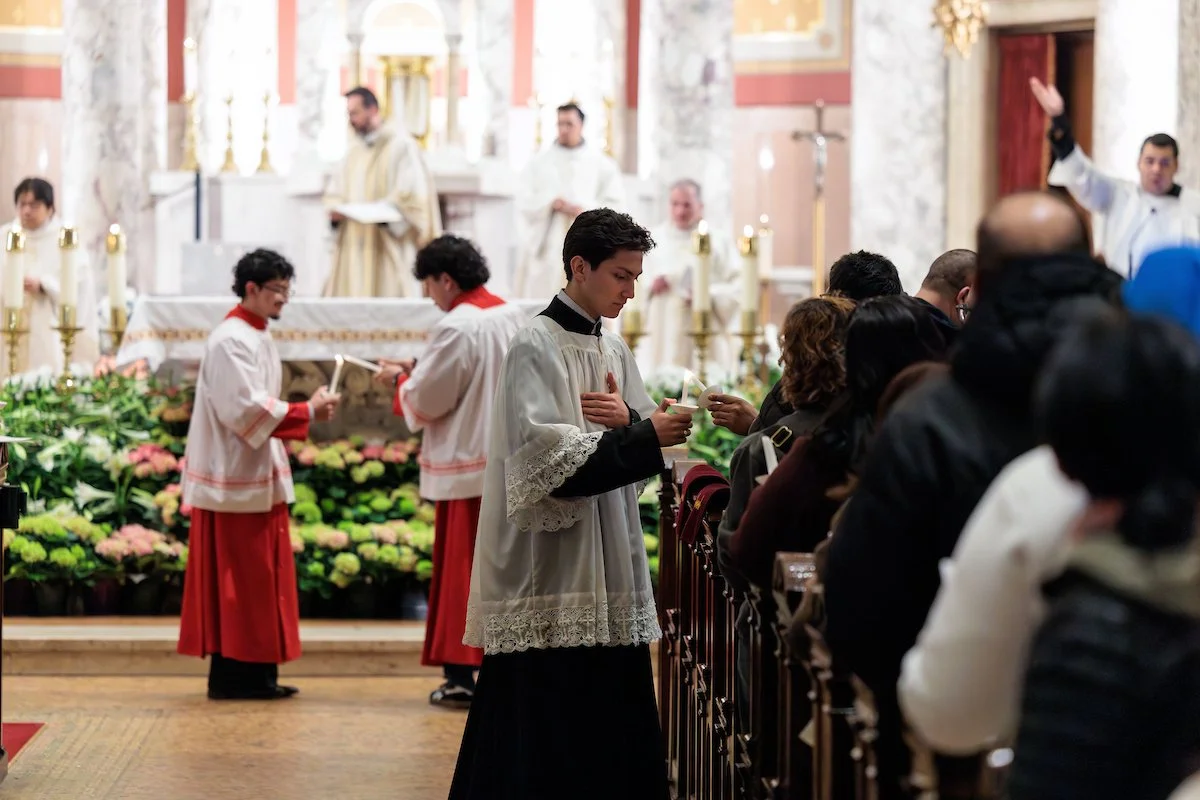 A young man in black and white clergy attire standing at the altar of a church, holding a candle and a card. A congregation of diverse people is seated nearby, with a priest and altar servers dressed in white and red robes visible in the background. The church interior is ornate with floral arrangements and candles.