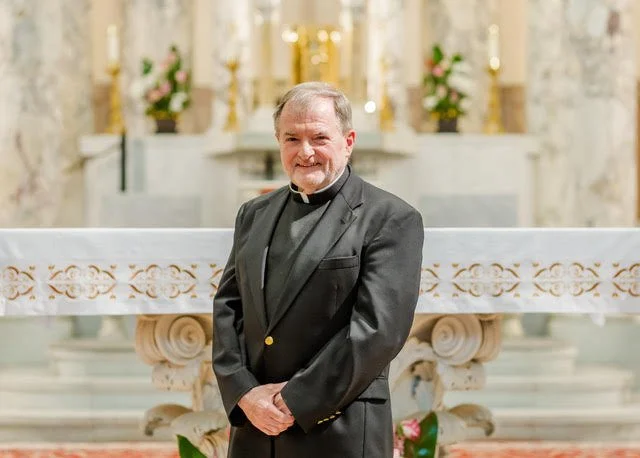 A priest in a black suit with a clerical collar standing in front of an altar in a church, smiling at the camera.