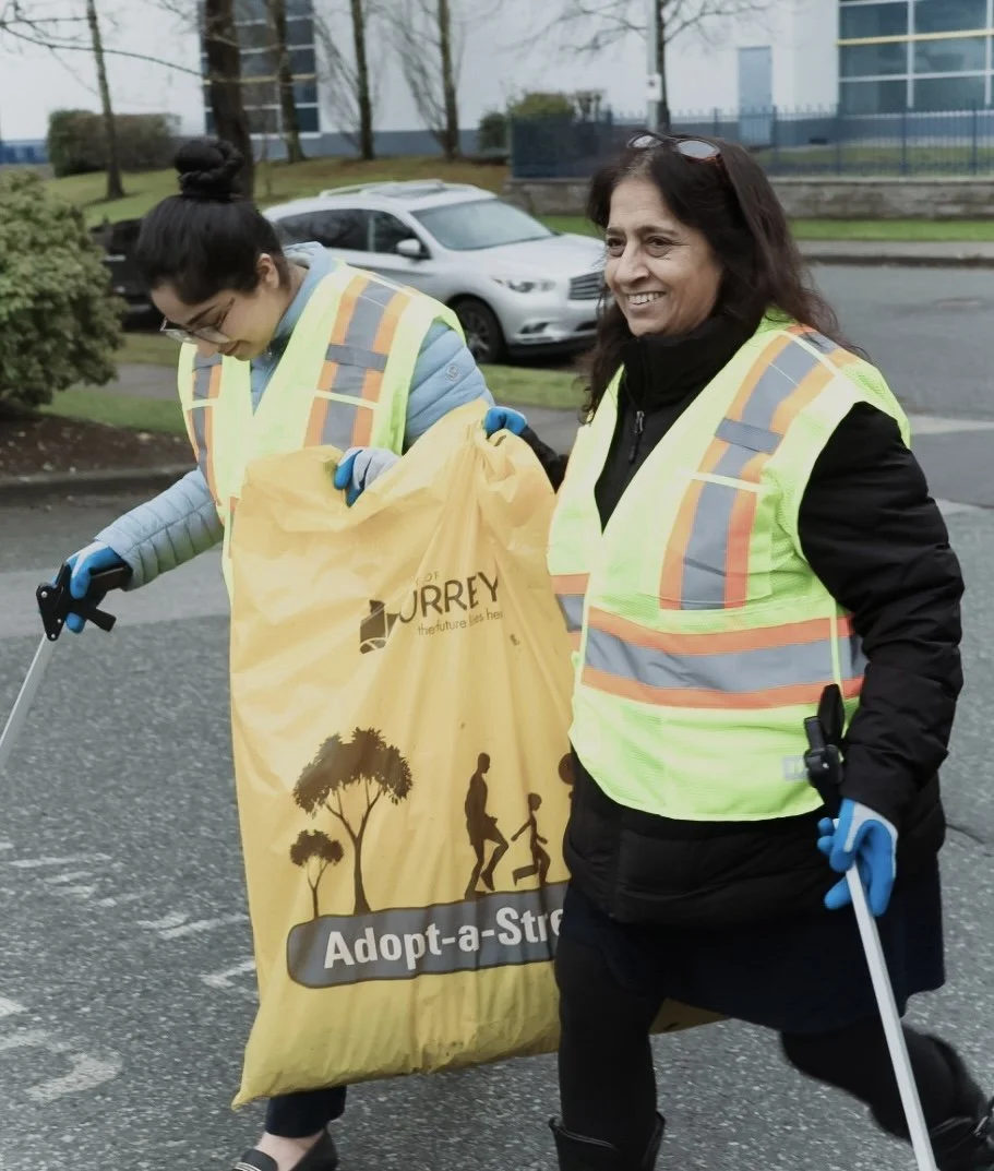 Two women in safety vests participating in a community cleanup, one holding a large yellow bag with an 'Adopt-a-Street' logo, walking on a street with parked cars and trees in the background. RBI Group. Community Participation. City of Surrey.