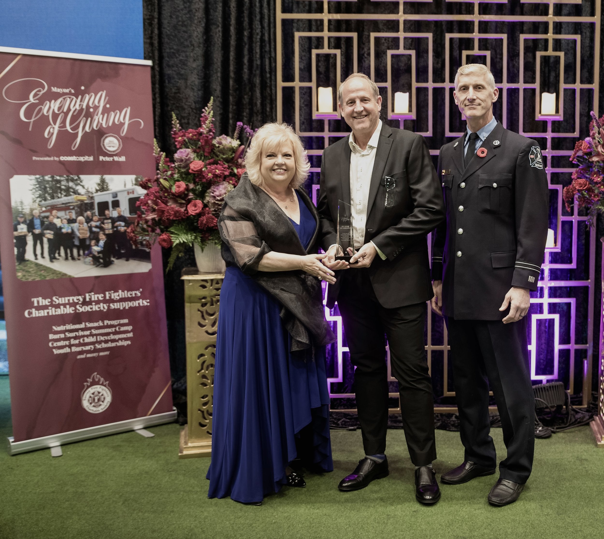 Three people standing in front of a decorated backdrop, two women and a man, holding an award. RBI Group. Community Participation. City of Surrey. Sukhi Rai. Mayor Brenda. Brenda Locke.