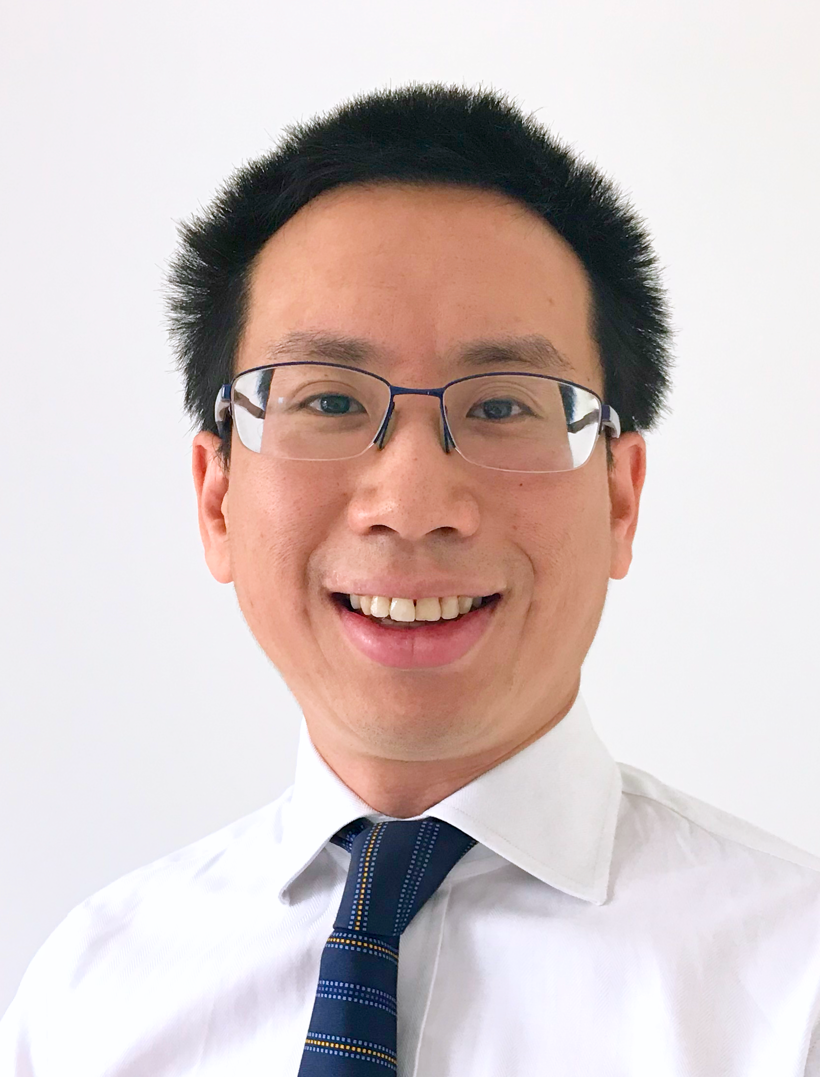 Photo of Lester Mak, a smiling man with black hair, glasses, wearing a white dress shirt and a dark striped tie, standing against a plain white background.
