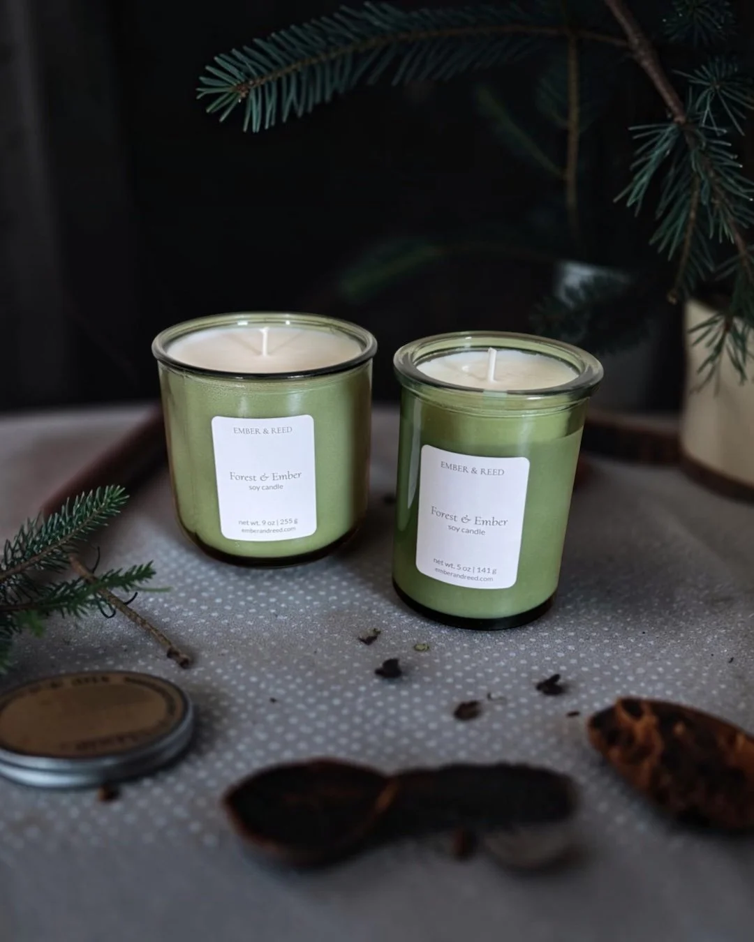 Two lit soy candles in a green recycled glass vessel with a label that reads 'Ember & Reed' and 'Forest & Ember,' placed on a textured surface with evergreens in the background.
