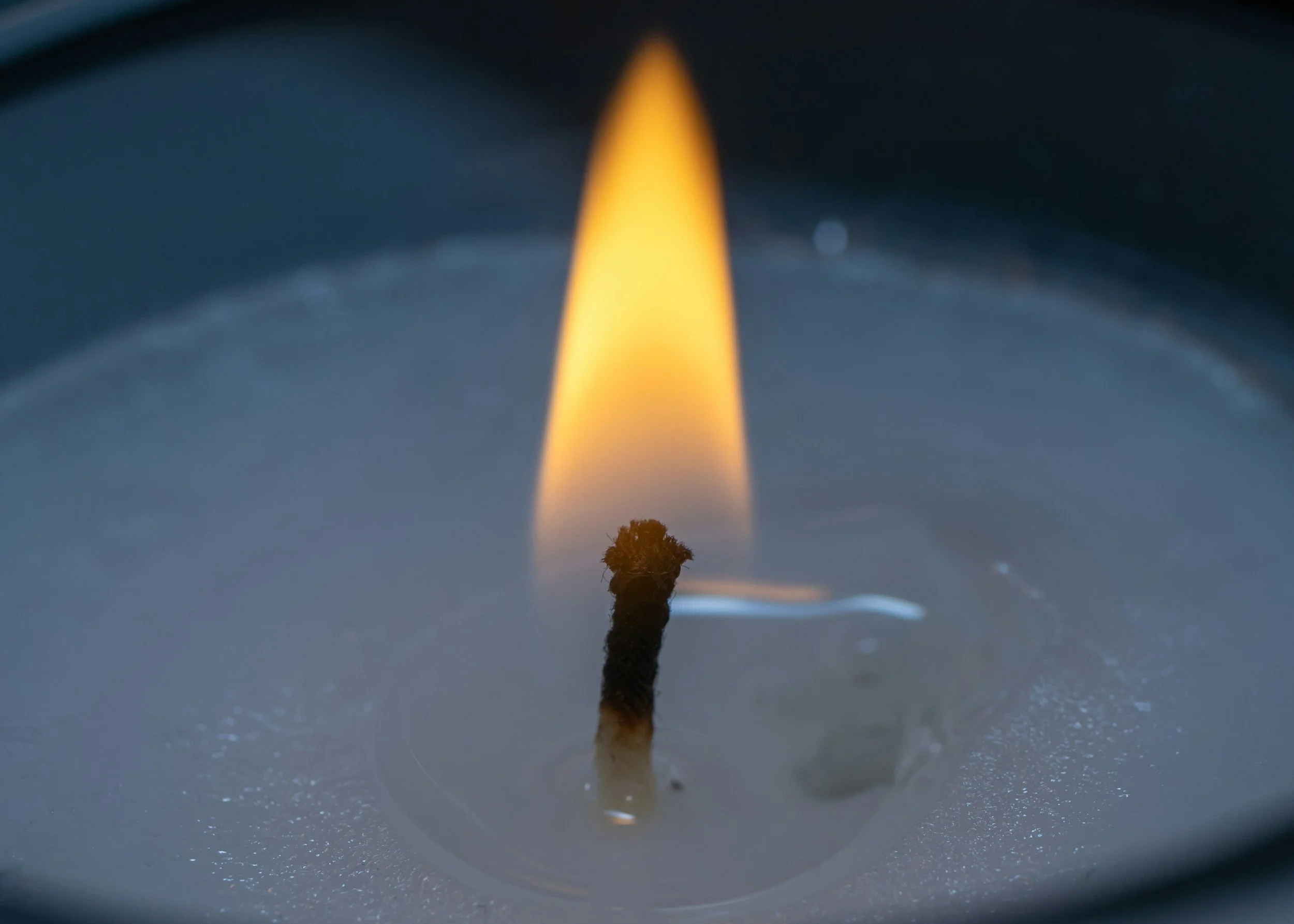Close-up of a lit candle with a yellow flame and blackened wick, melted wax surrounding the wick.