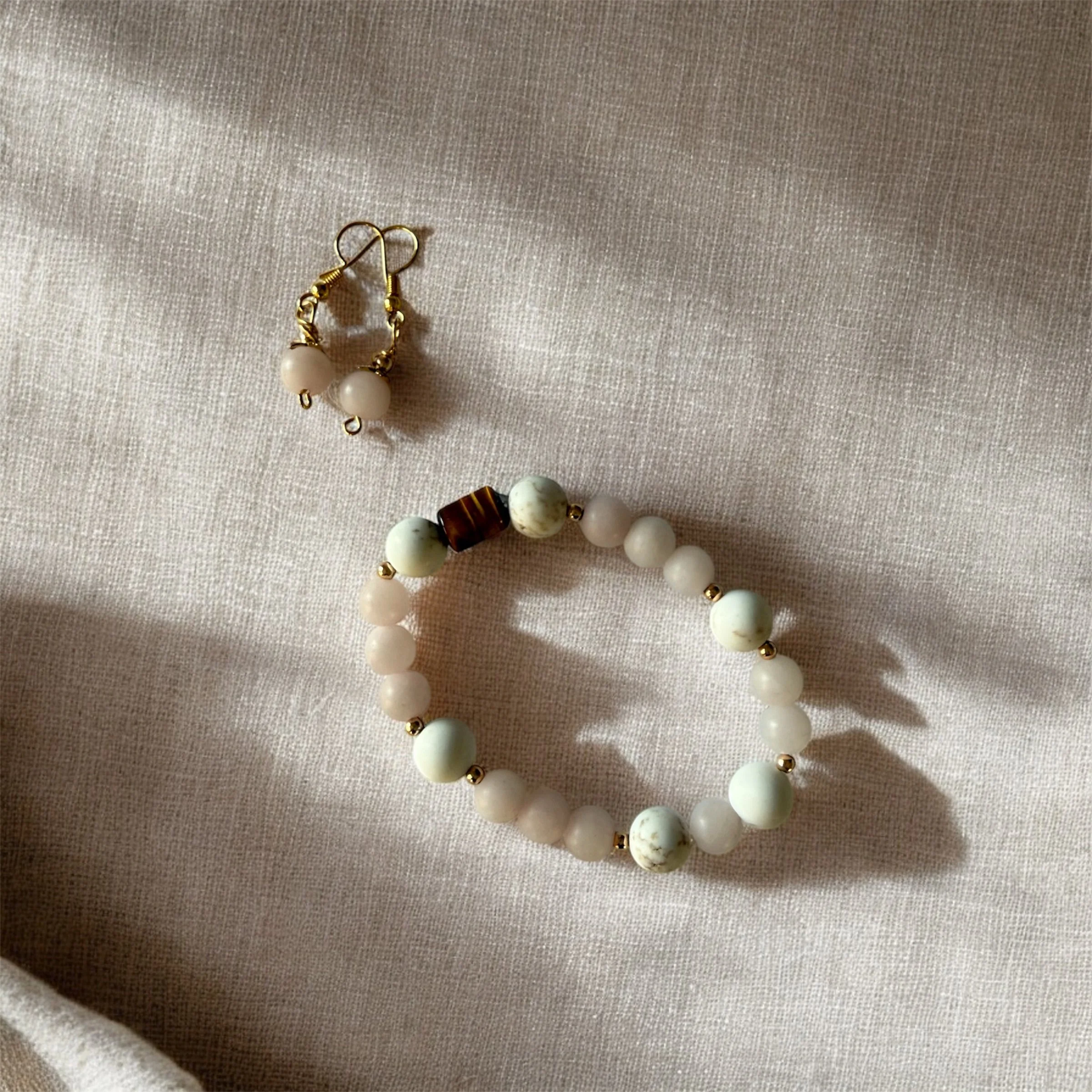 A pair of blush quartz earrings and a tiger's eye and blush quartz beaded bracelet on a light fabric surface.