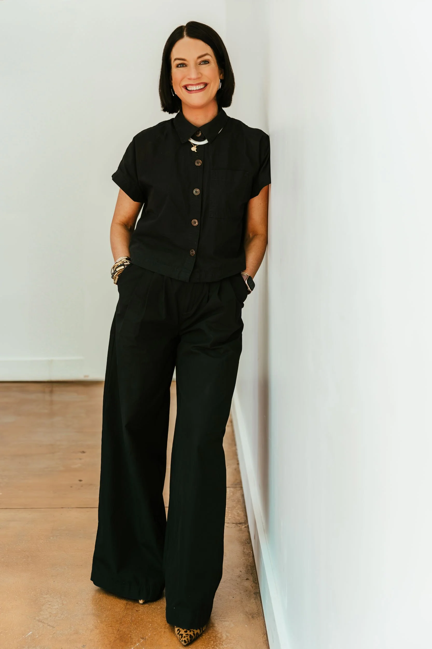 Reluctantly Styled founder Teresa Dillon, wearing a black short-sleeve button-up shirt, black wide-leg pants, smiling and leaning against a white wall.