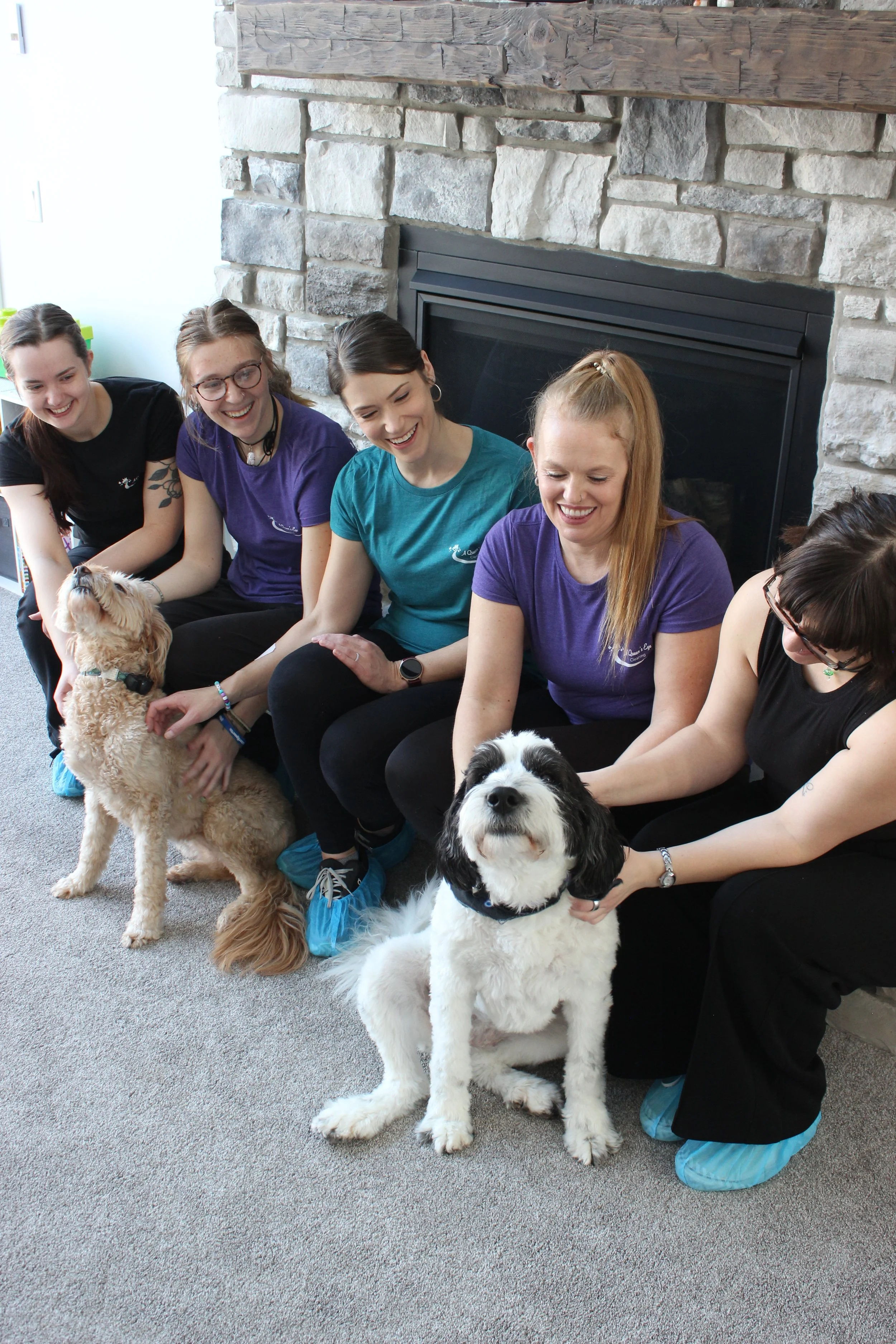 Five women sitting in front of a fireplace, each with a dog, smiling and interacting with the dogs.