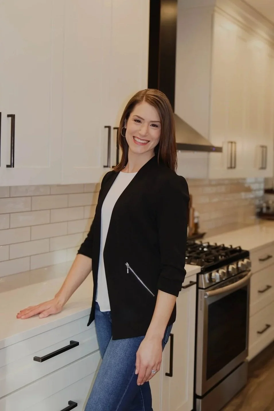 Smiling woman with brown hair in a black blazer, white top, and blue jeans standing in a modern kitchen with white cabinets and stainless steel stove.